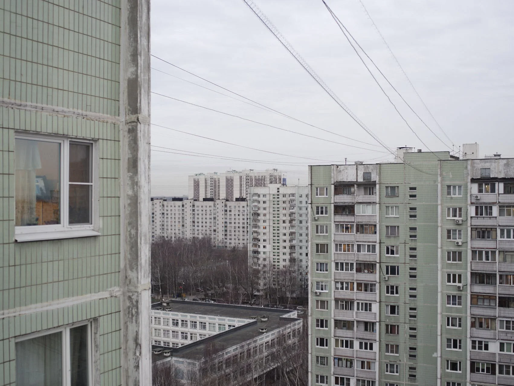 View of several high-rise apartment buildings with electric wires overhead, on a cloudy day, with some trees and a street below.