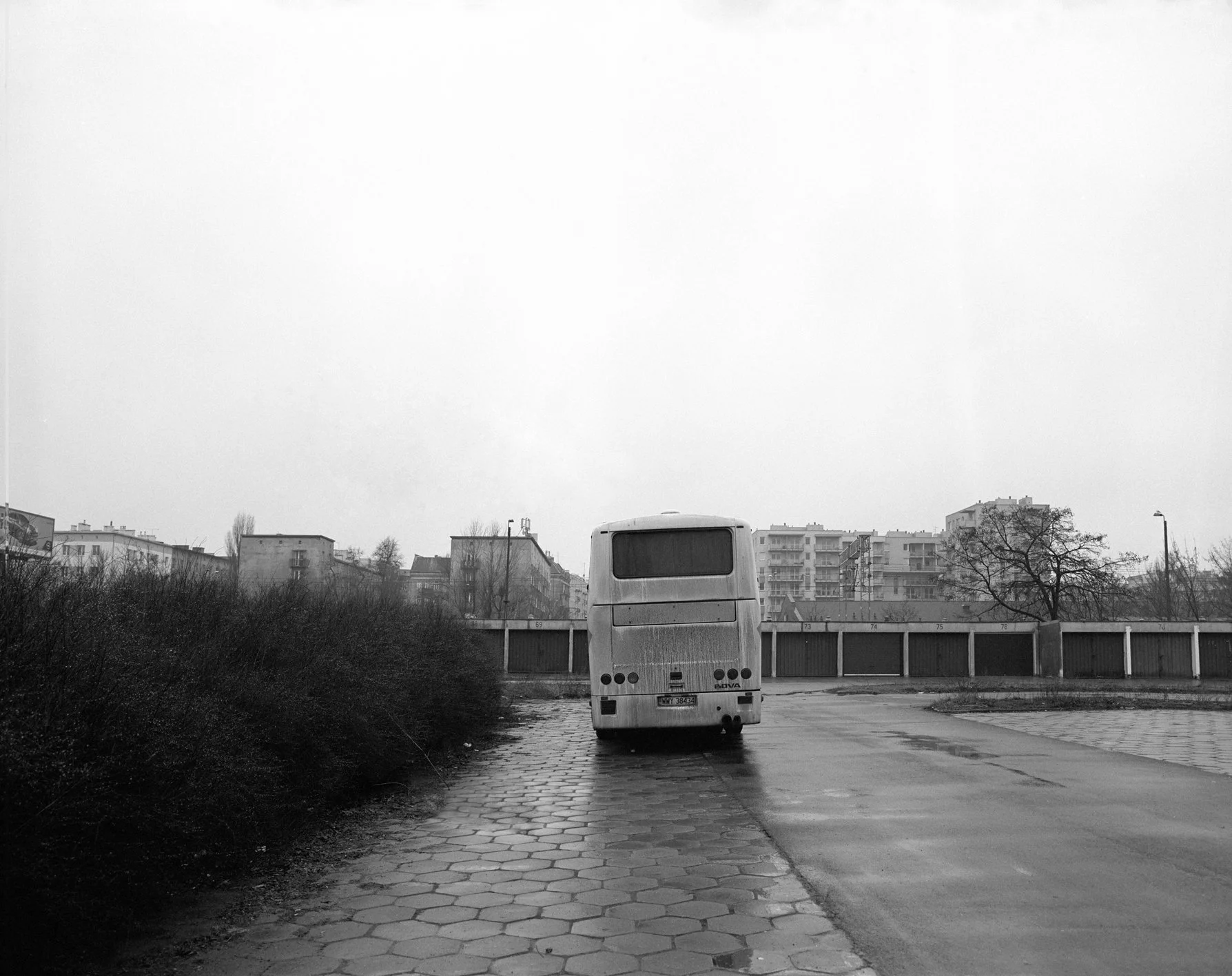 Black and white photo of a bus parked on a wet sidewalk near bushes and buildings in the background on a cloudy day.