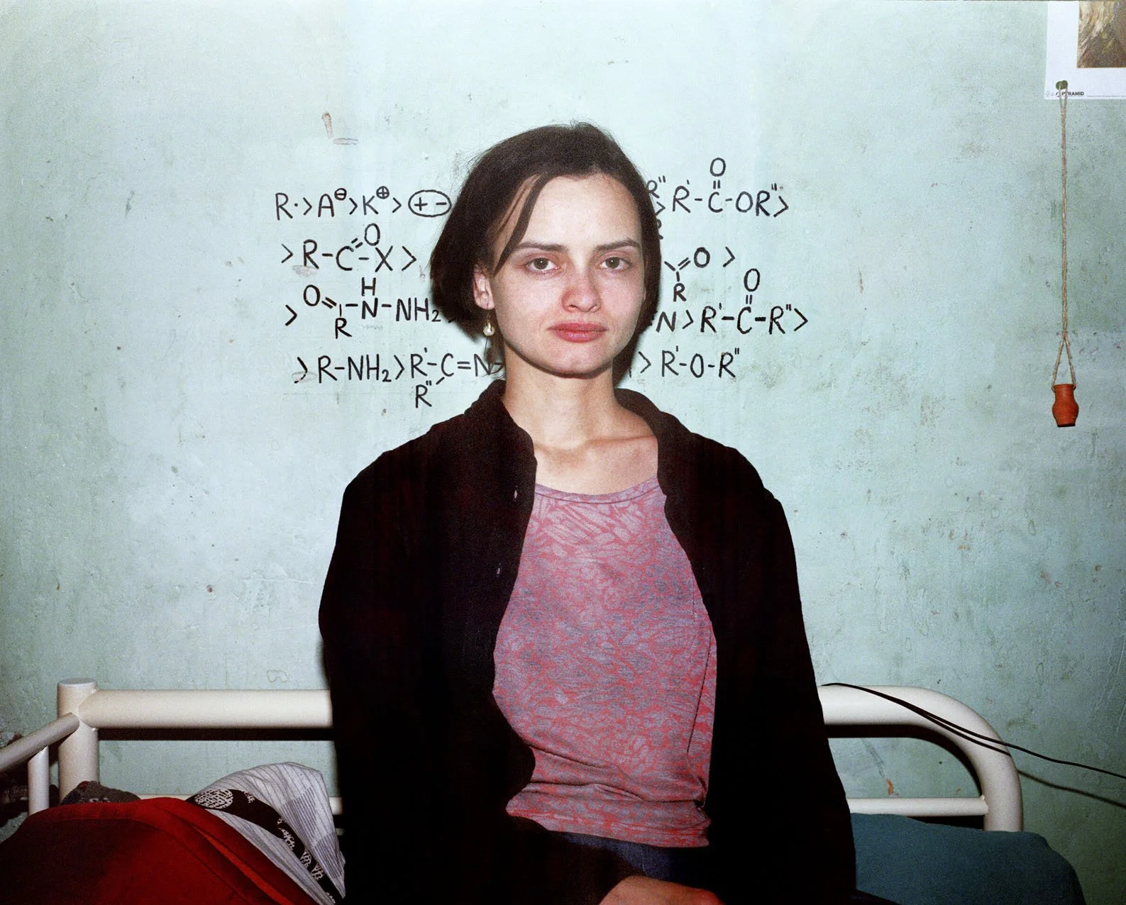 A young woman with short dark hair sitting on a bed in front of a wall with chemical diagrams written on it, wearing a pink shirt and a black cardigan.