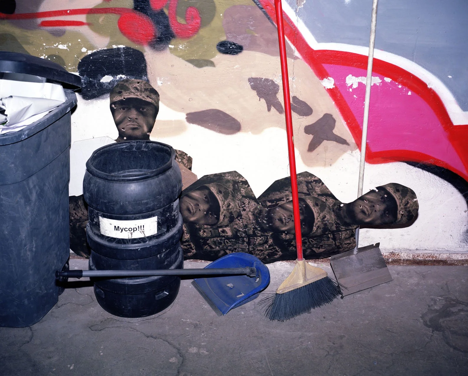 Street art mural with soldiers in camouflage uniforms, some lying down, with a background of colorful graffiti. In front of the mural are a black trash bin labeled "Mycop!!!", a barrel, a dustpan, a broom, and other cleaning tools.