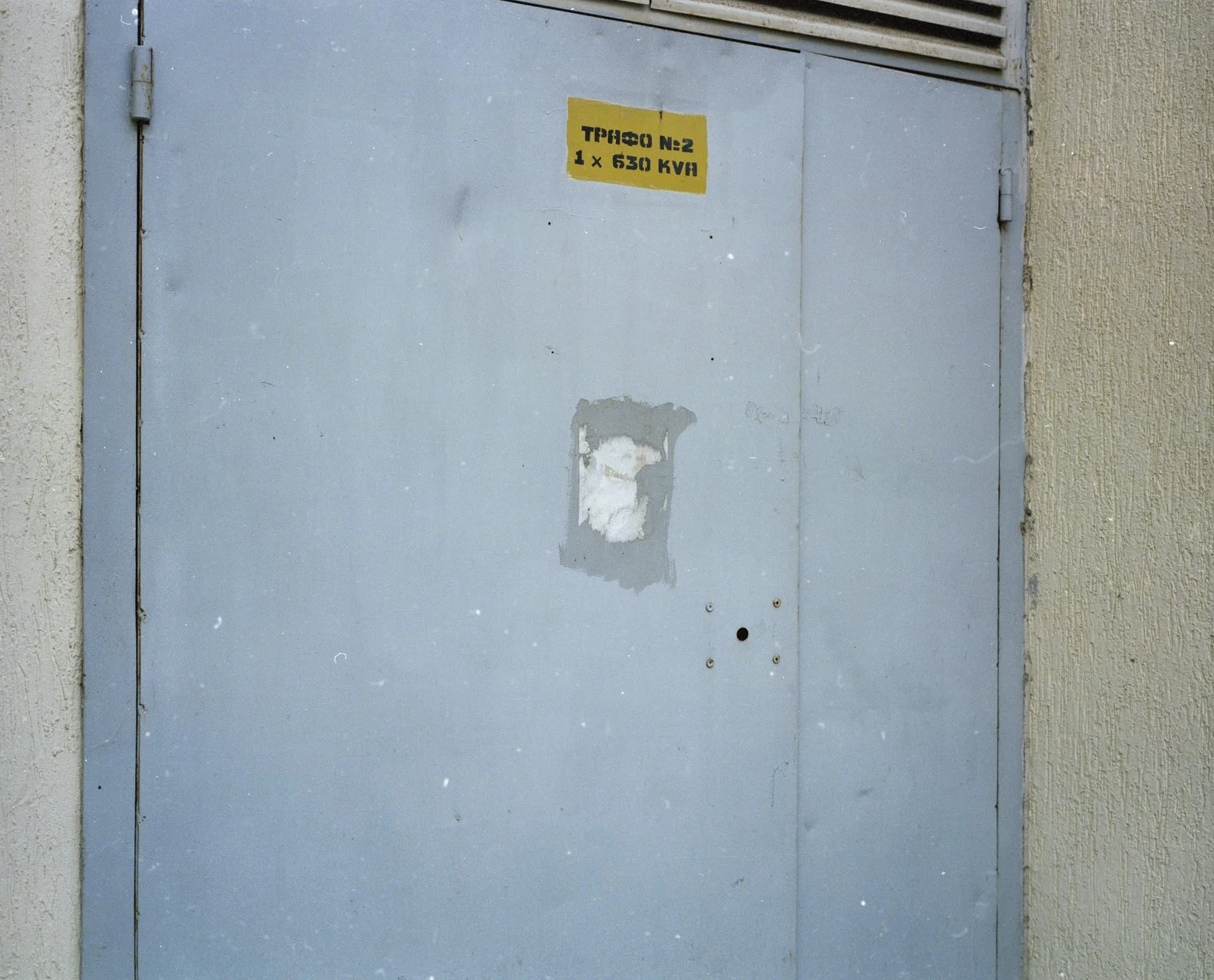 A gray metal utility door with a partially worn yellow caution sticker on the top center that has black text. The door is surrounded by a beige wall with a textured surface. There are small holes and a patch of peeling paint on the door.
