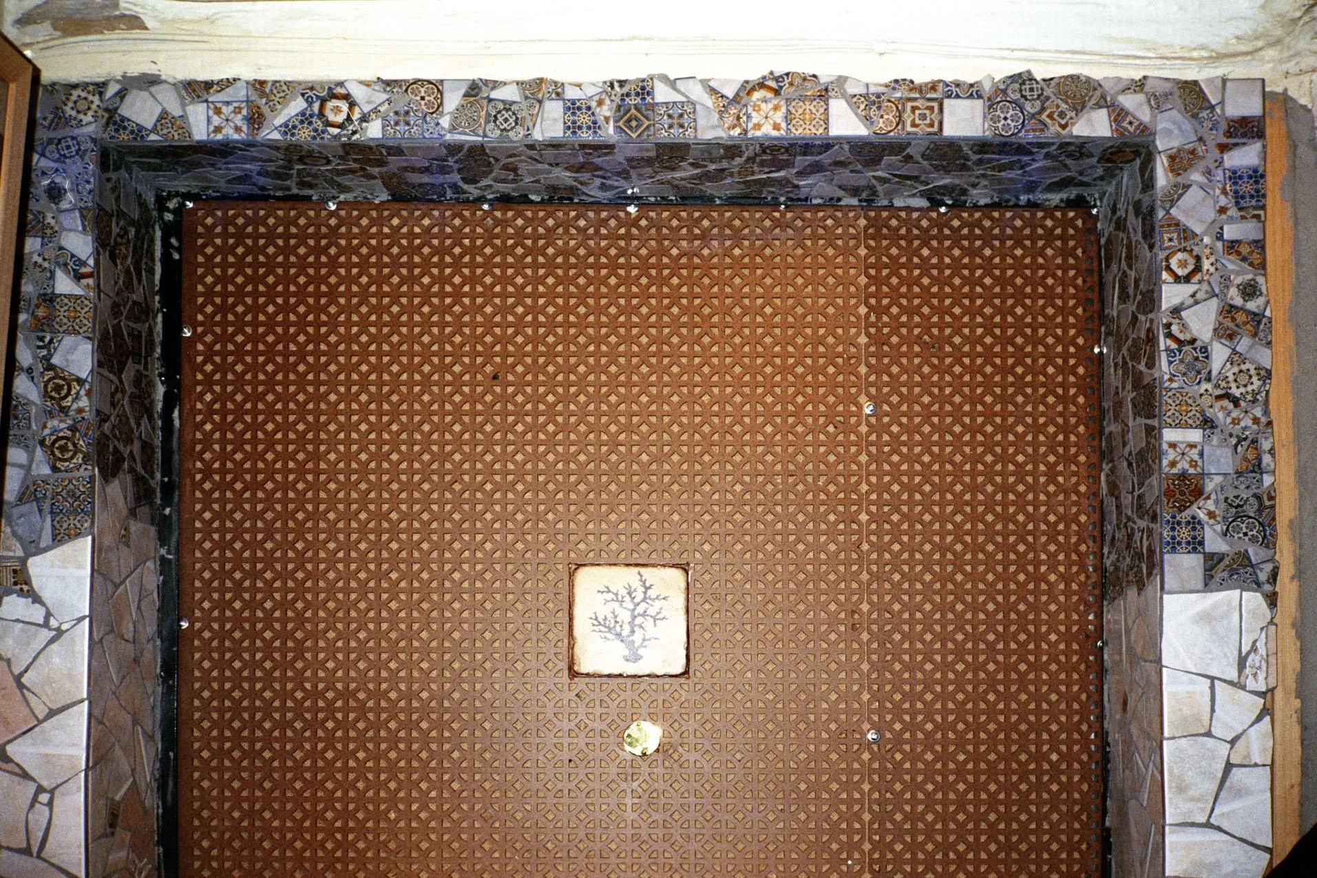 A square, brown metal cover with a geometric pattern on the ground, surrounded by a border of irregularly shaped tiles with colorful, intricate designs.