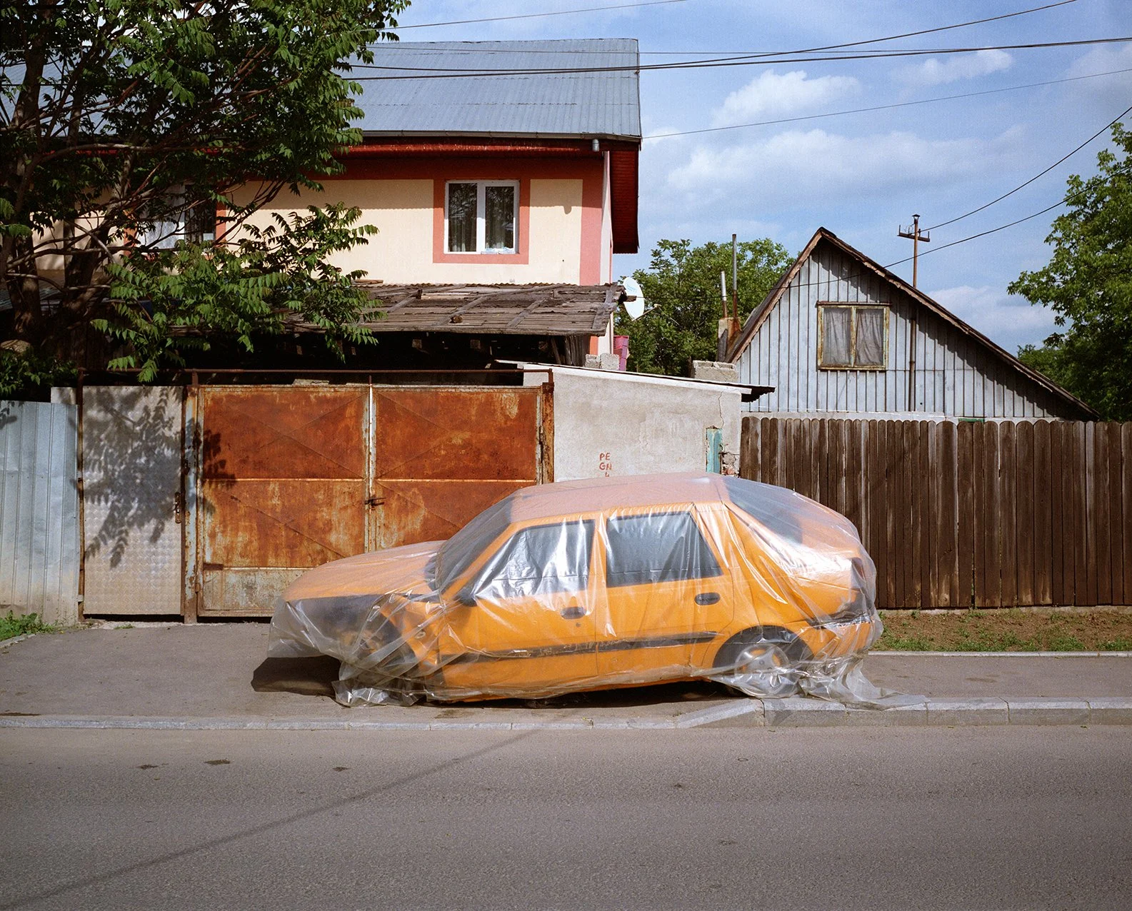 An orange car covered in a clear plastic sheet parked on the side of the street in front of a rusty gate and wooden fence, with houses and trees in the background.