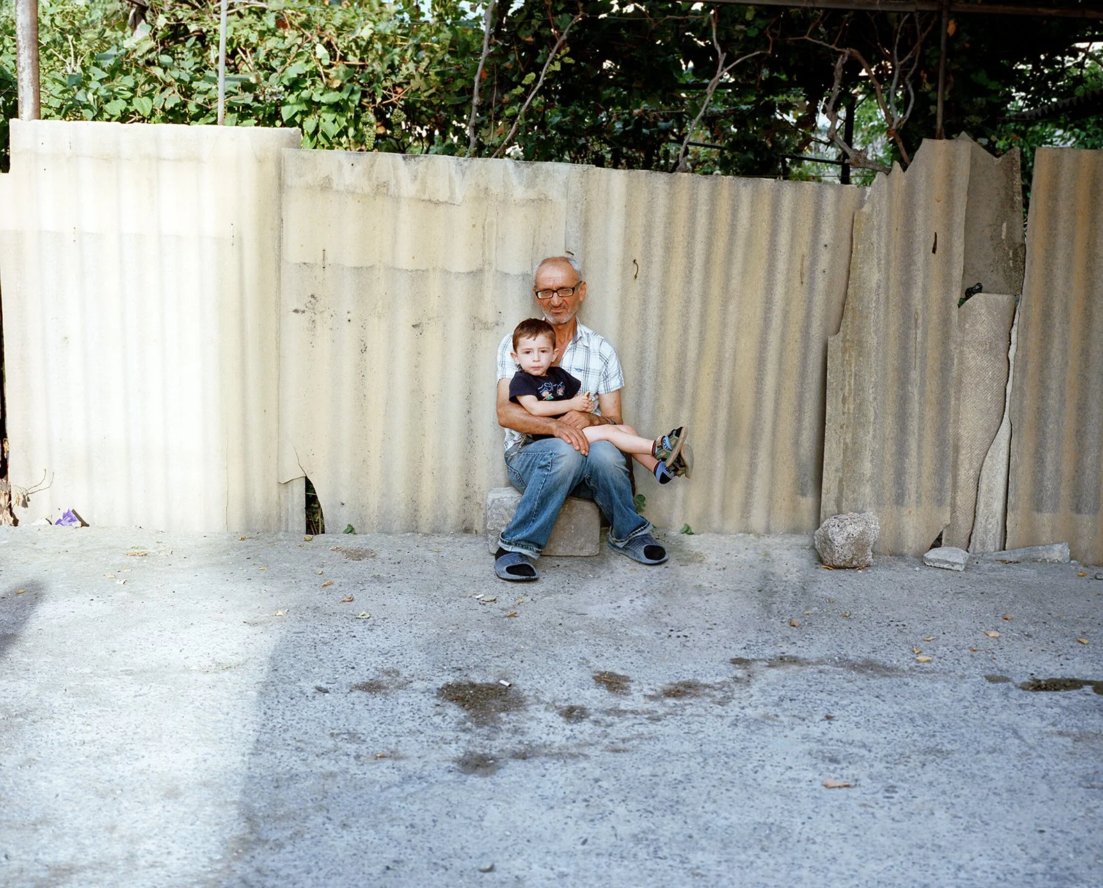 An older man and a young boy sitting together on a concrete bench in front of a corrugated metal fence with green foliage behind it.