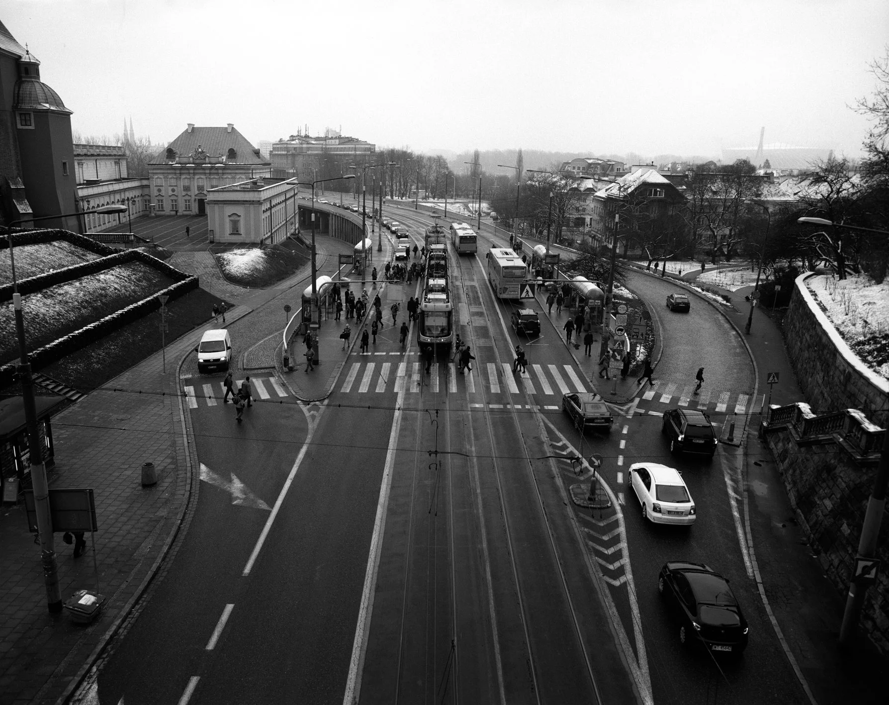 Urban street scene with cars and buses waiting at a tram stop, pedestrians crossing the street, and snow on the ground in a city with historic buildings.
