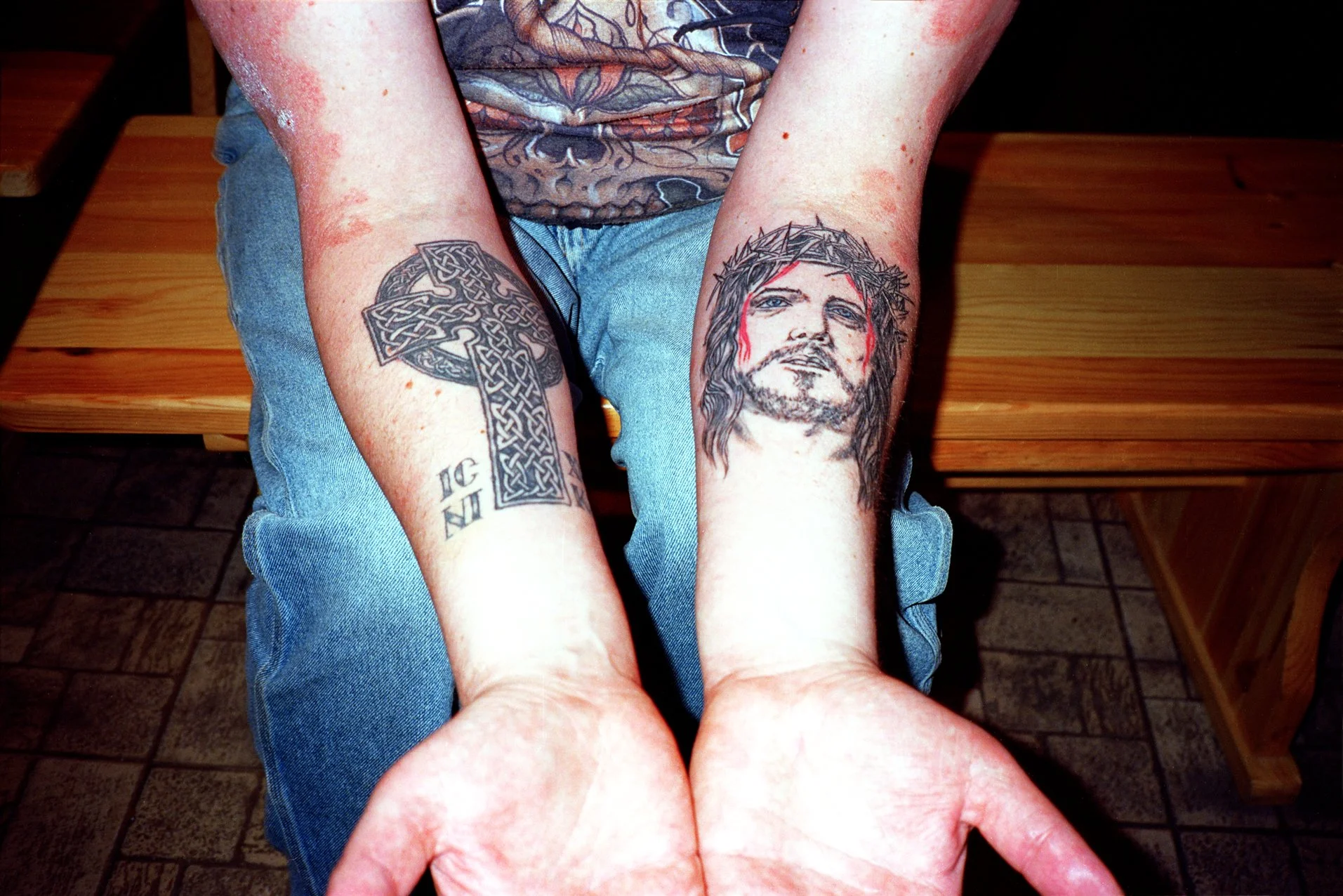 Close-up of a person's arms resting on their knees, showcasing two tattoos: a Celtic cross on the left arm and a portrait of Jesus Christ with a crown of thorns and red tears on the right arm. The person is wearing a T-shirt with a dragon graphic and denim shorts, seated on a wooden bench.