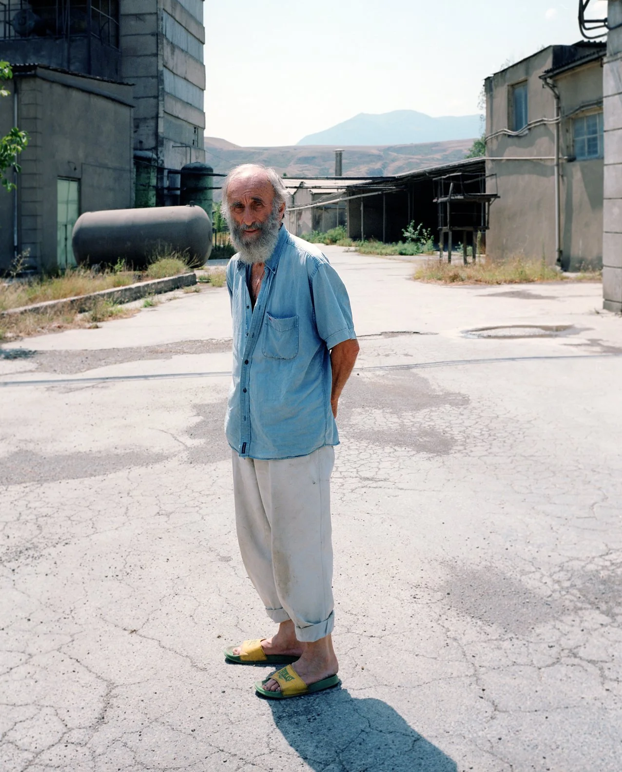 An elderly man with a white beard in casual clothing stands in an empty, cracked street in an industrial area with worn buildings and a landscape of mountains in the background.