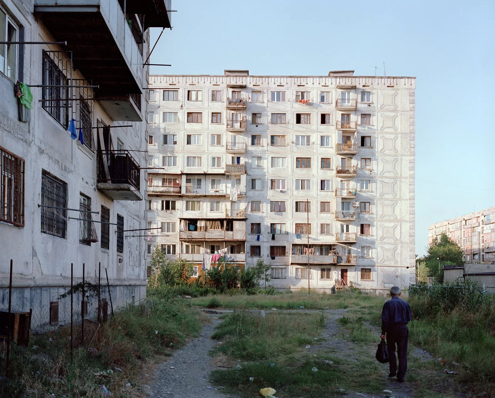 A man walking along a dirt path in front of tall, weathered apartment buildings with balconies, laundry hanging, and overgrown grass.