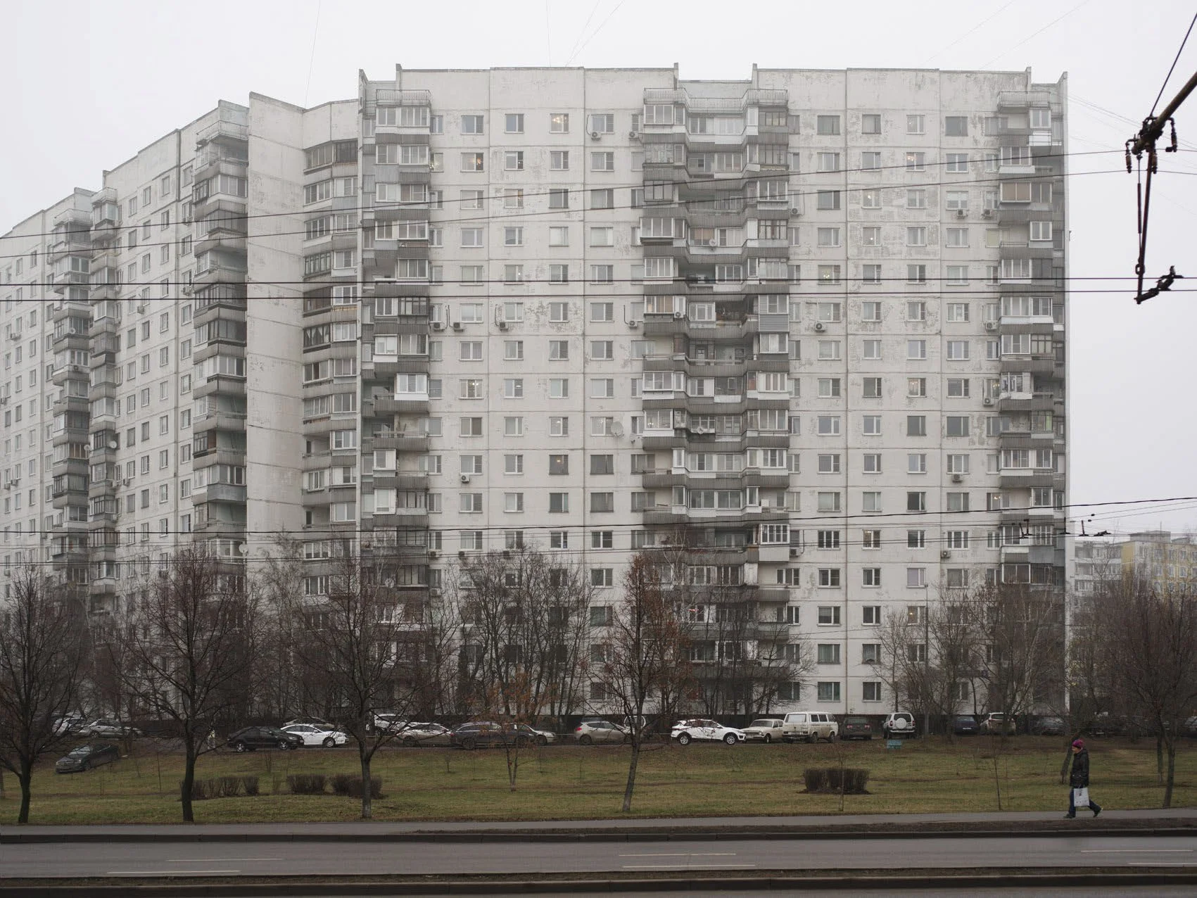 A large, gray, multi-story residential apartment building with many balconies, surrounded by leafless trees and a parking lot with cars. A person walks on the sidewalk in the foreground on a cloudy day.