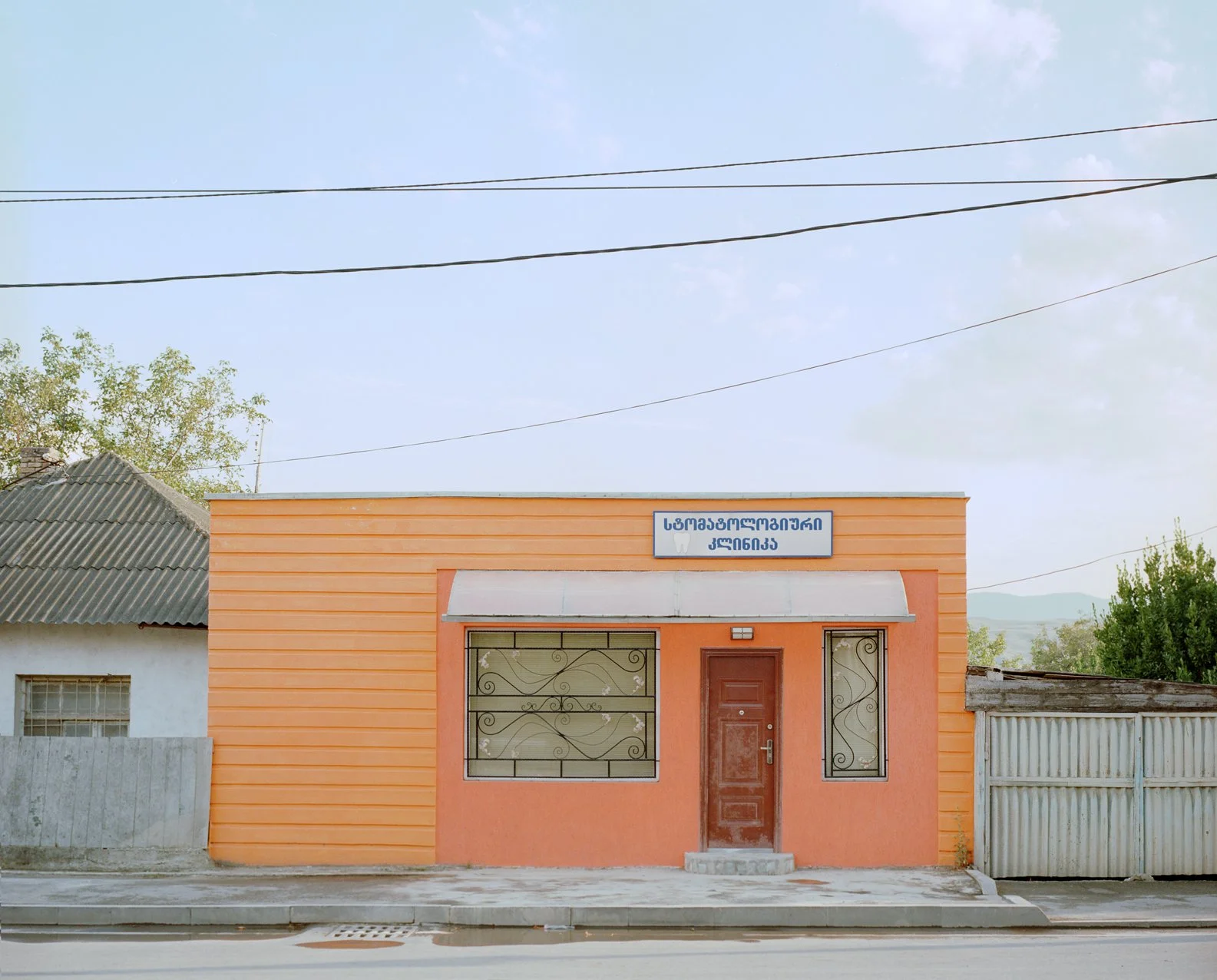 Small orange building with a sign in Georgian above the door and a decorative front door with wrought iron bars on the windows, situated on a street with a concrete sidewalk and adjacent structures, under a partly cloudy sky.