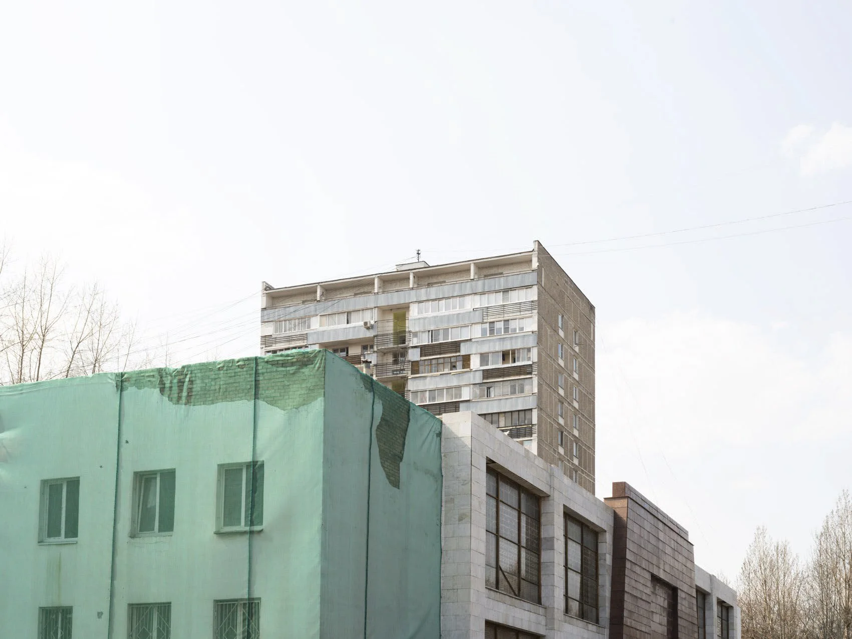A high-rise apartment building with multiple balconies and windows, partially obscured by an adjacent smaller building wrapped in green construction netting.