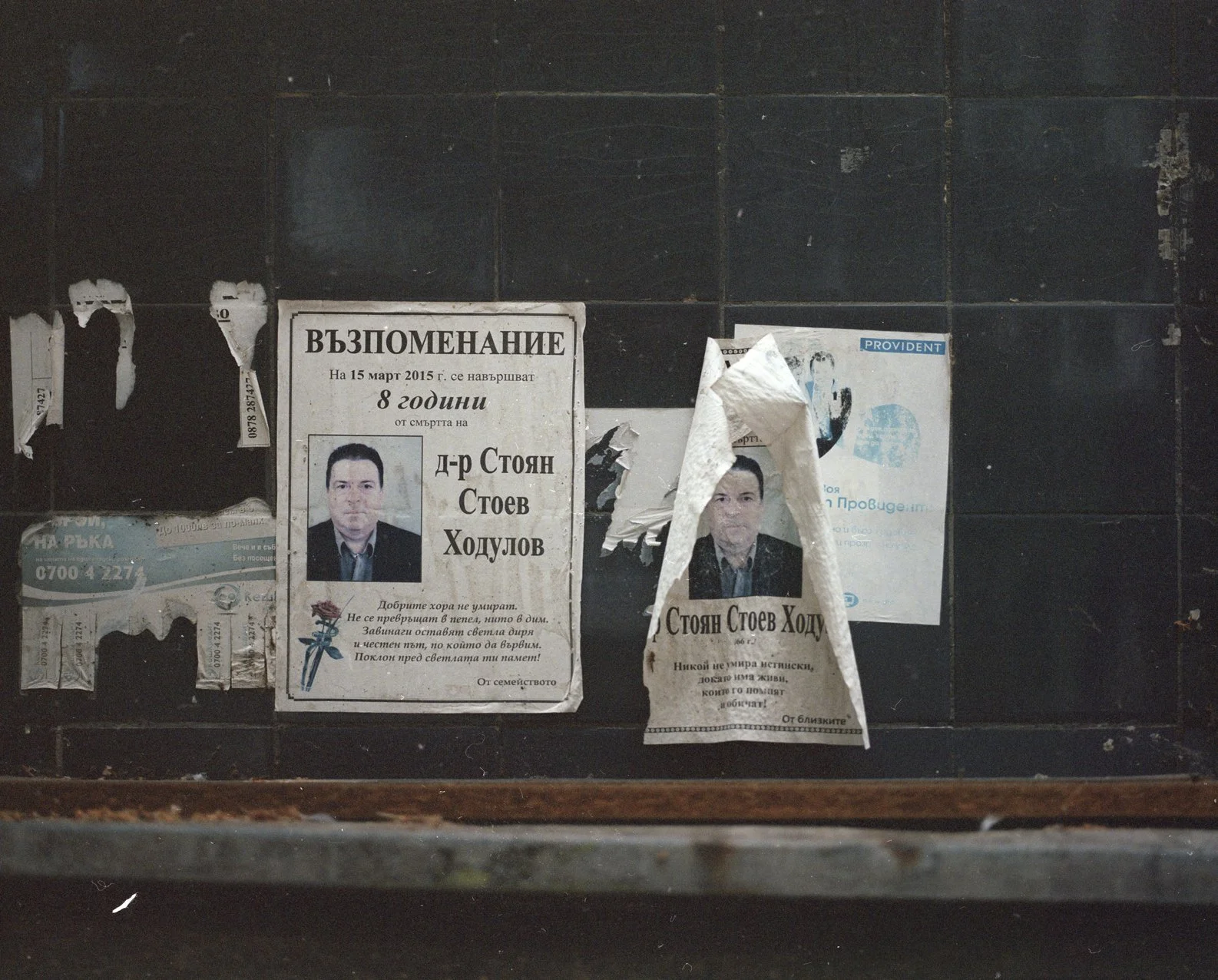 Four torn posters on a black tiled wall, one of which features a portrait of a man and text in Bulgarian, with some peeling and crumpled papers.