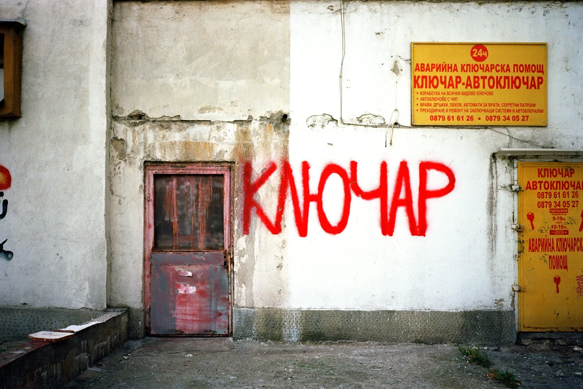 Graffiti on a white wall with a small pink door, reading 'ключар' in red spray paint. Two yellow signs with red text in a language other than English are mounted on the wall to the right of the graffiti.