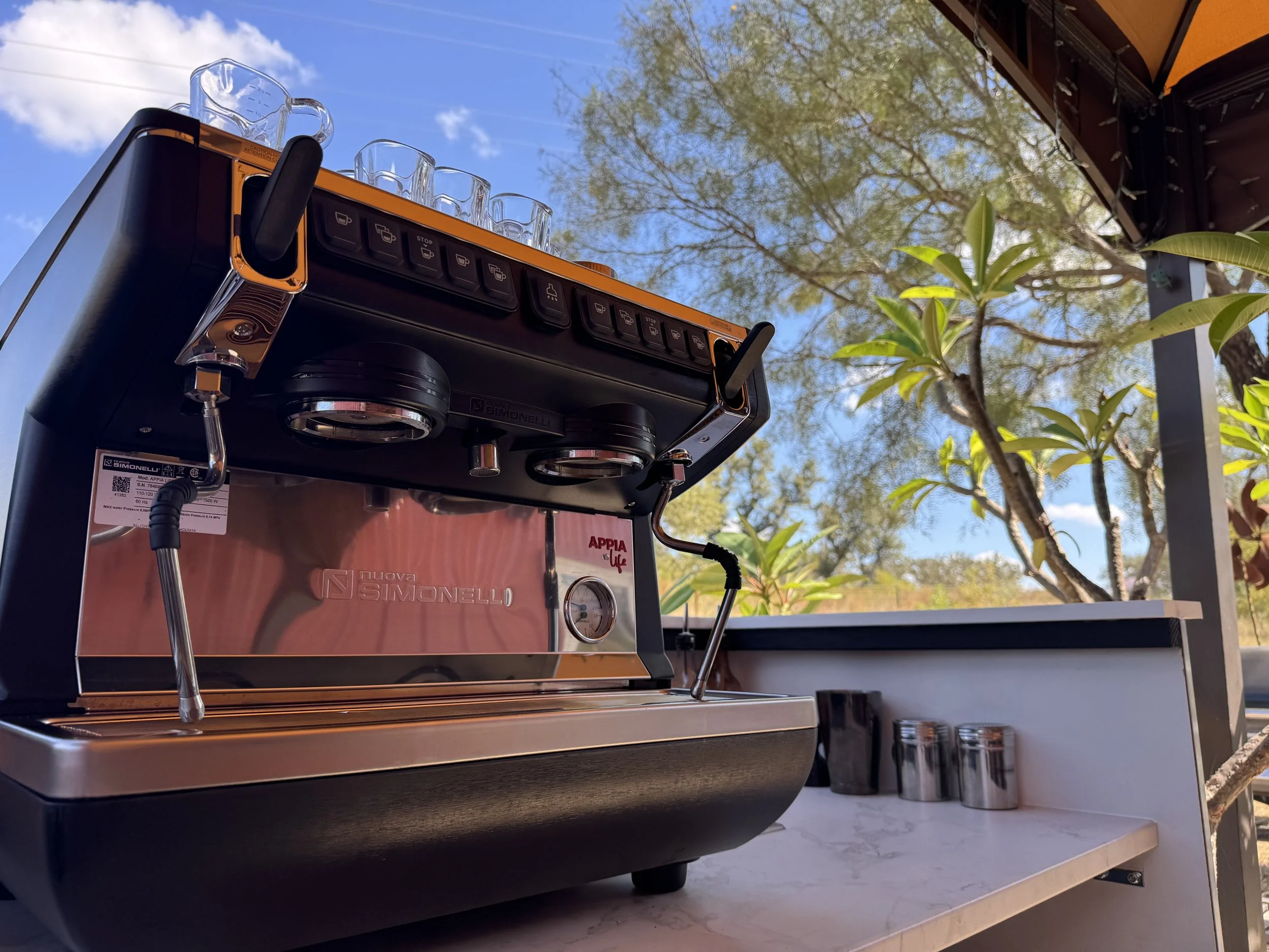 Close-up of a black and silver espresso machine on a white marble countertop outdoors, with a tree and blue sky in the background.