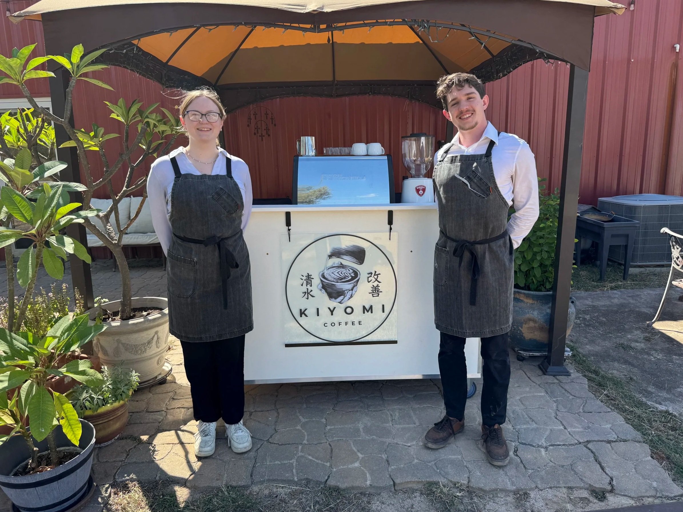 Two people standing outdoors behind a small coffee stand with a sign reading "Kiyomi Coffee". Both are wearing aprons and smiling. The stand is under a canopy, with potted plants around.
