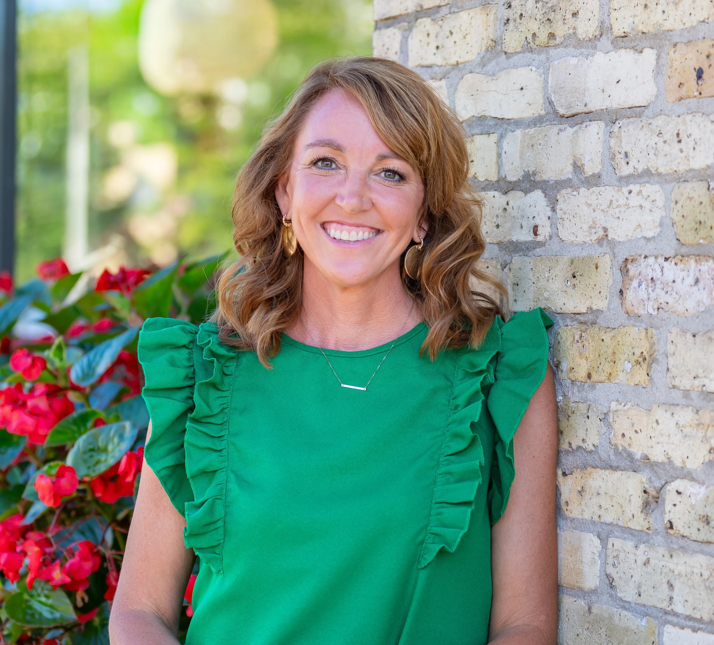 A woman with shoulder-length wavy brown hair and blue eyes leaning against a brick wall outdoors, wearing a green top with ruffled sleeves and gold jewelry, smiling at the camera with flowers and greenery in the background.