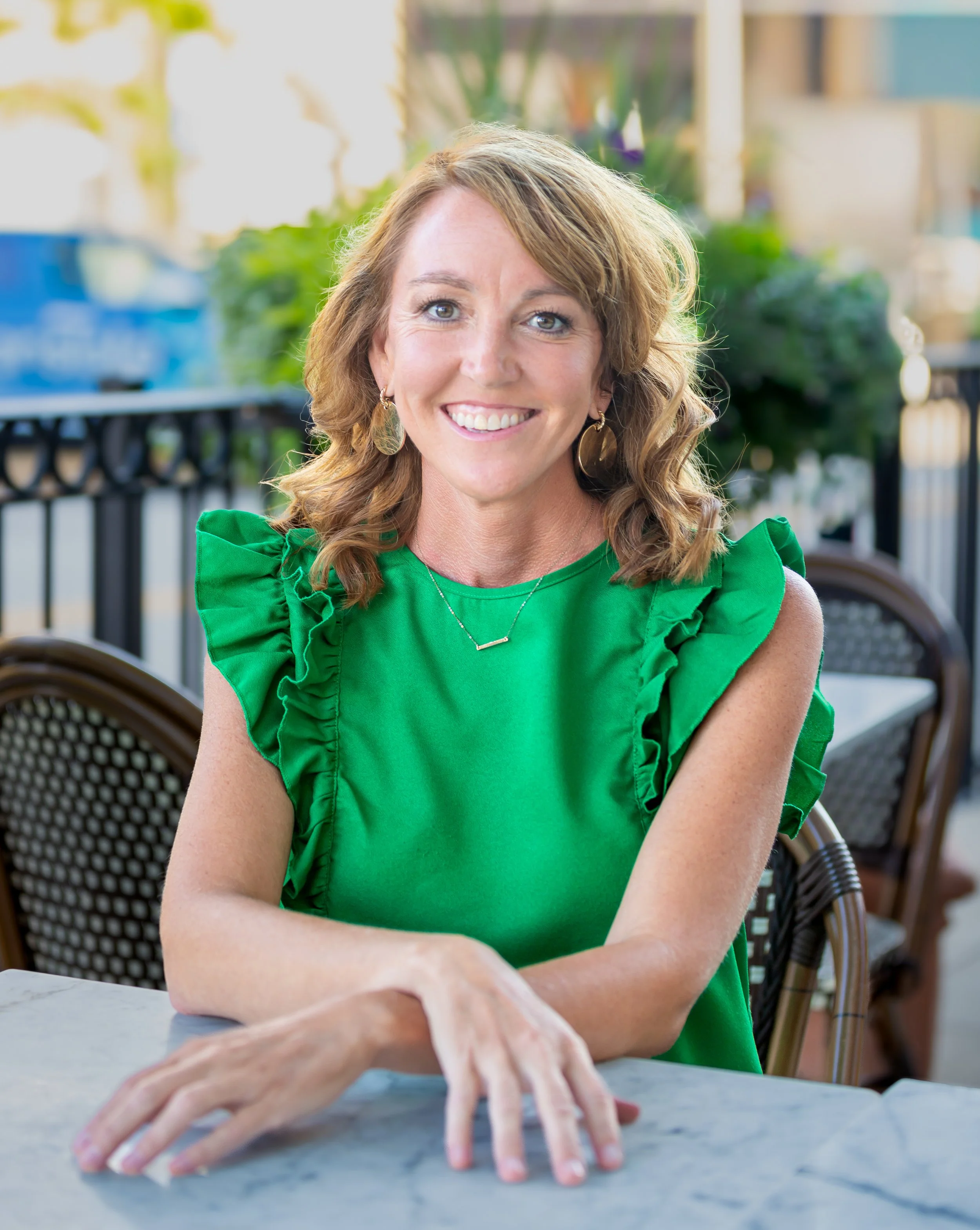 A woman with shoulder-length curly light brown hair, wearing a bright green sleeveless top with ruffled sleeves, and gold earrings, sitting at an outdoor cafe table with a marble top, smiling at the camera.