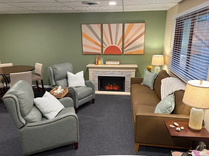 Cozy living room with green wall, fireplace, and modern decor. Features two light blue armchairs, a brown sofa, and a small round dining table with white chairs. Decor includes pillows, lamps, and artwork of a sun with rays on the wall.