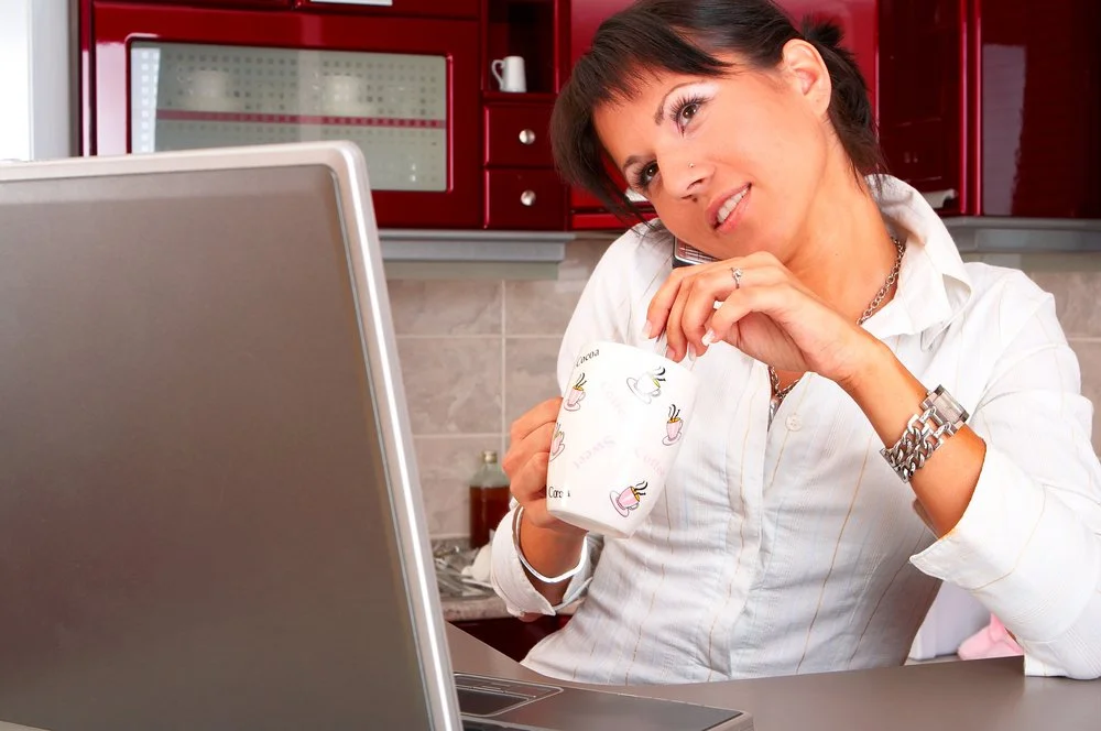 A woman in a red-cabinet kitchen talks on the phone while stirring a hot drink at her laptop, balancing a multi-tasking work-from-home routine.