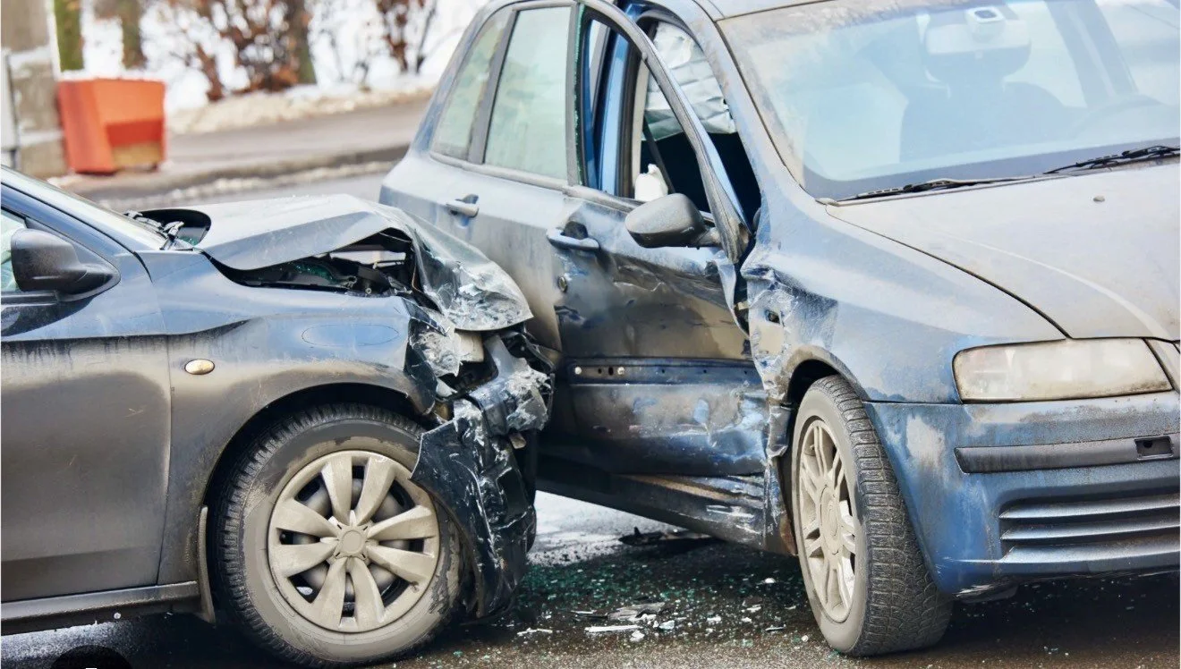 A realistic photograph of a T-bone car accident showing a dark sedan with a crushed front end impacted into the side of a blue vehicle, illustrating the severe mechanical forces that lead to whiplash and soft tissue trauma.