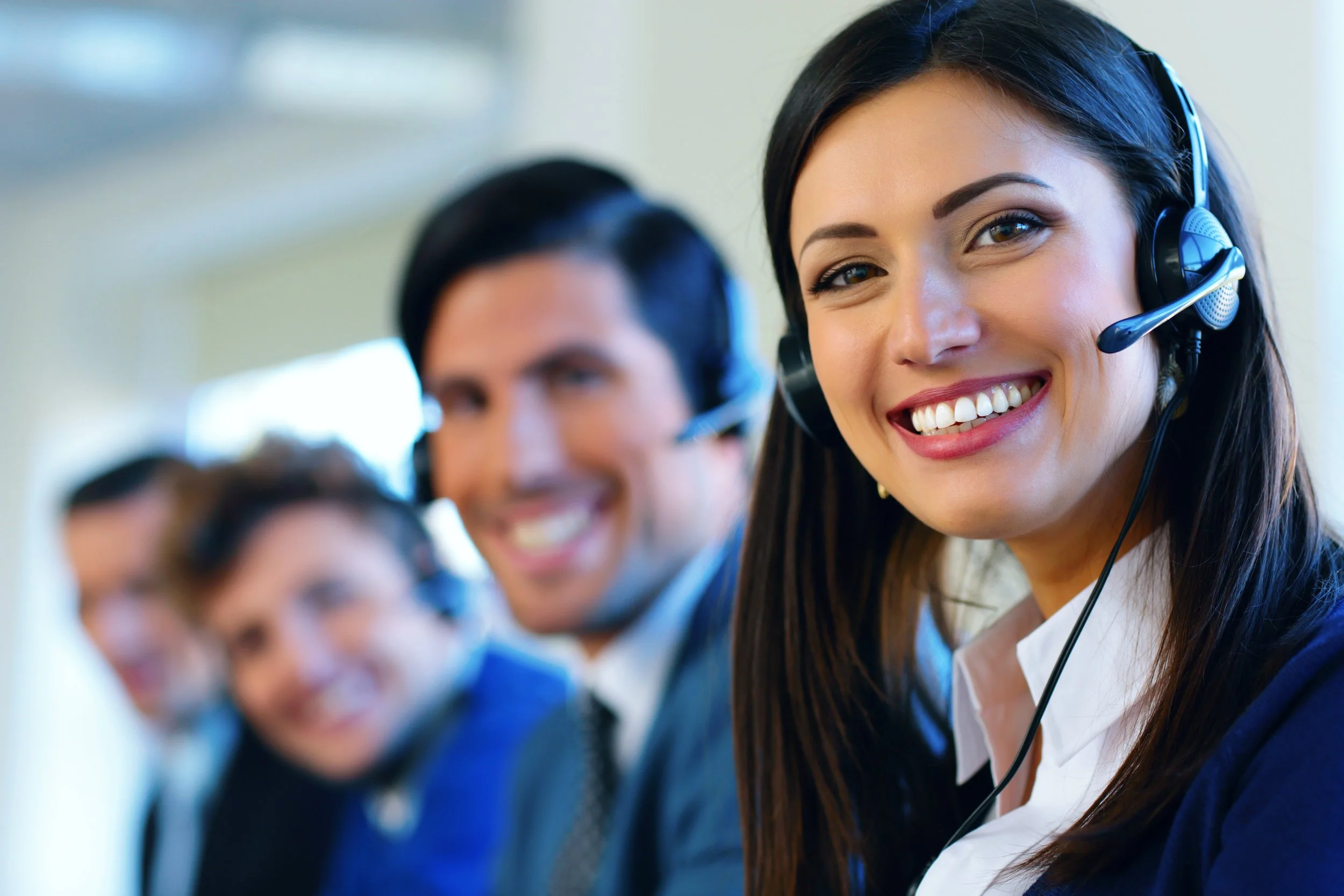 A close-up of a smiling woman with long dark hair wearing a professional black headset with a morcophone suggesting a professional call center or customer service environment.