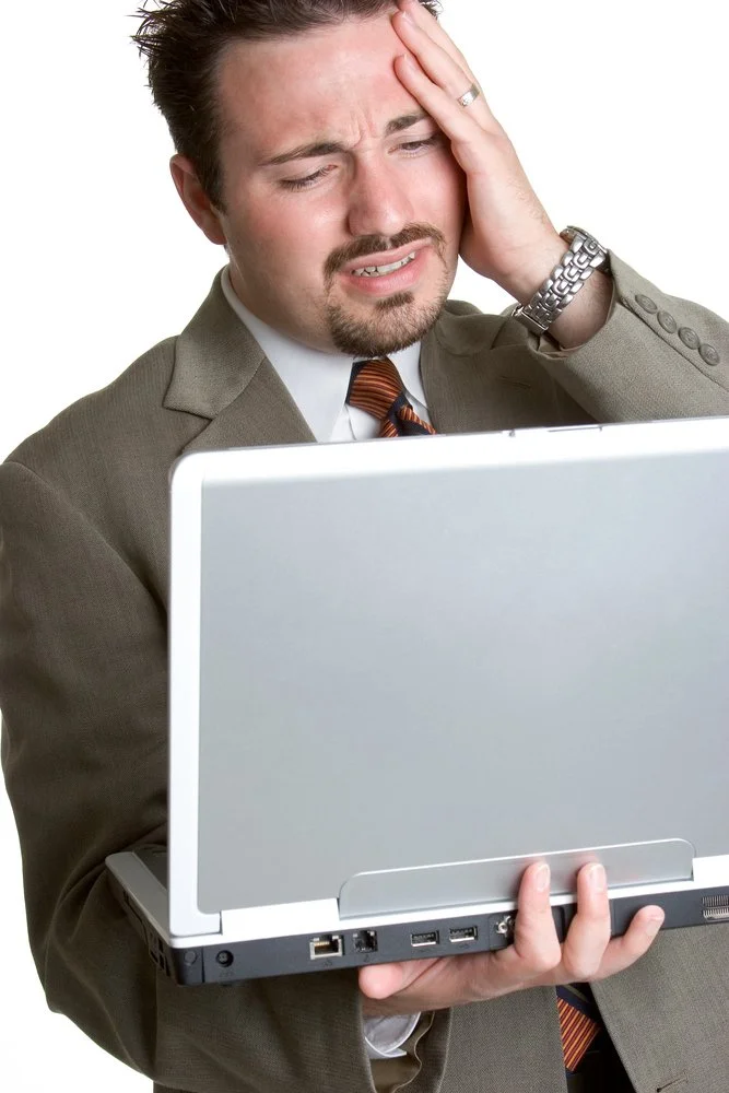 A man in professional attire holding his head in discomfort while looking at a laptop, representing post-concussion headaches and radiating shoulder pain often caused by whiplash and mild traumatic brain injuries (mTBI).