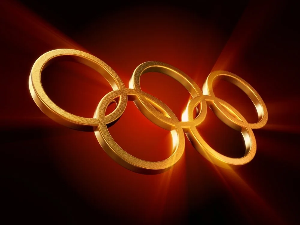 Gemini said  A three-dimensional render of five interlocking golden rings, based on the Olympic symbol, set against a glowing, deep red background with radial light beams bursting from behind.