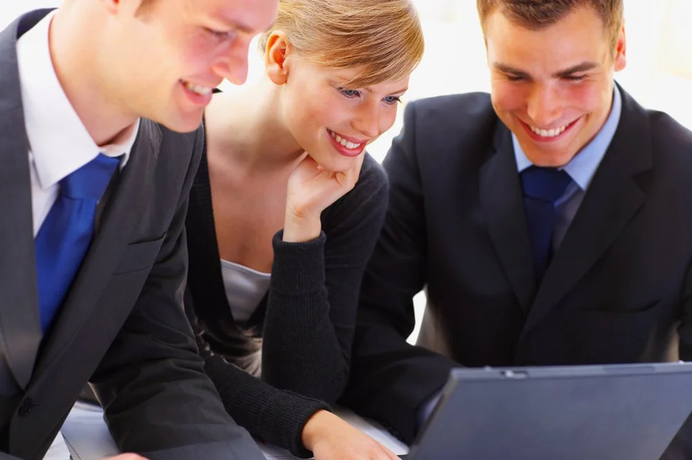A closeup of three collegues looking and smiling at a laptop closely in a business enviroment. A woment in a blue blaser sits between two men whom are wearing gray suits.