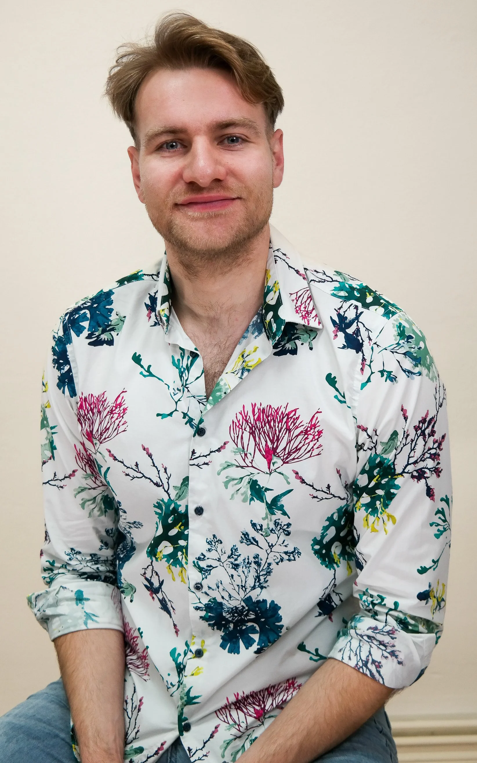 Ein junger Mann mit lockigem, hellbraunem Haar trägt ein weißes Hemd mit buntem Blumenmuster. Er lächelt leicht vor einem neutralen, hellen Hintergrund.