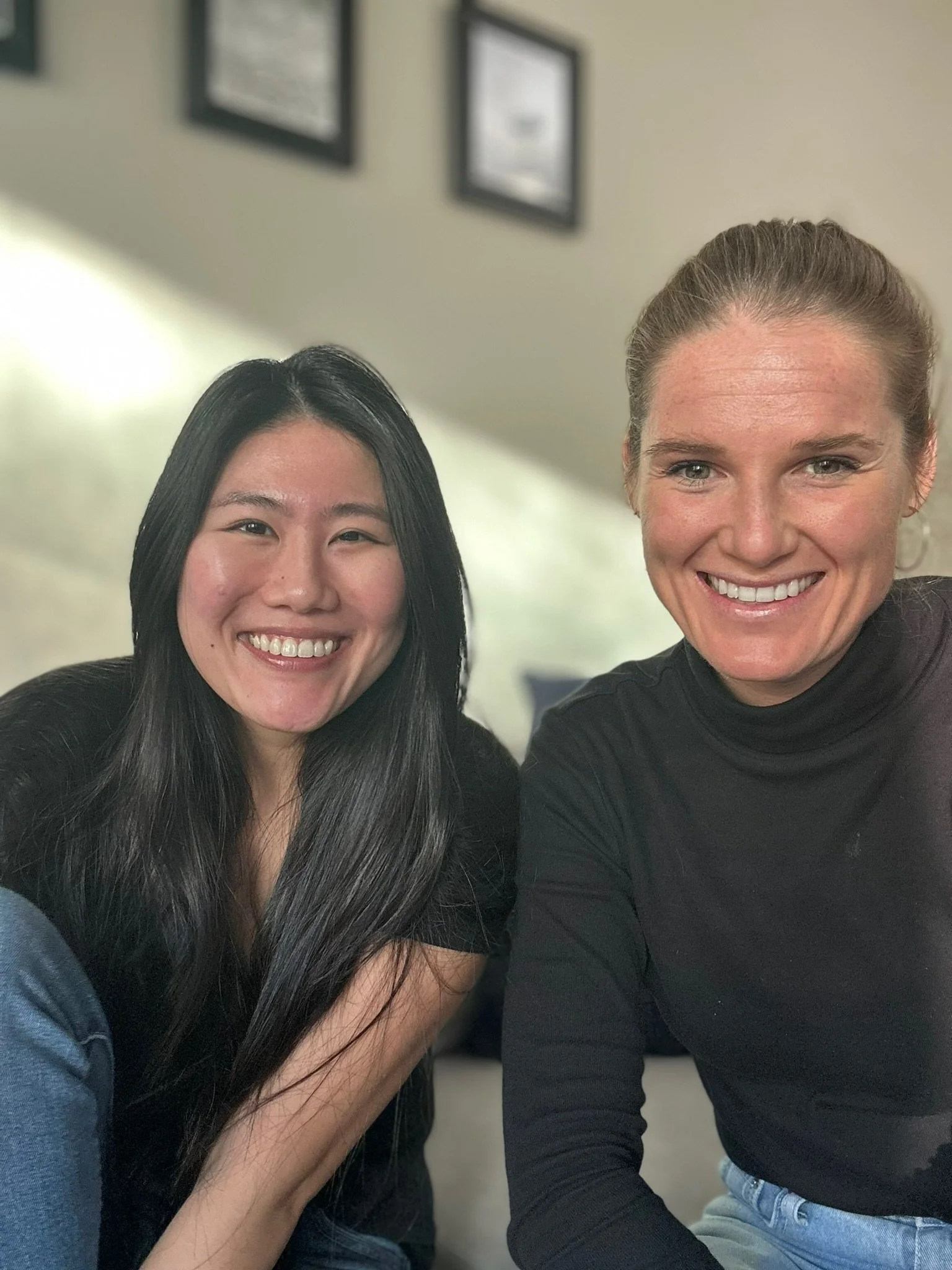 Two women smiling and sitting close together indoors, with framed pictures on the wall behind them.
