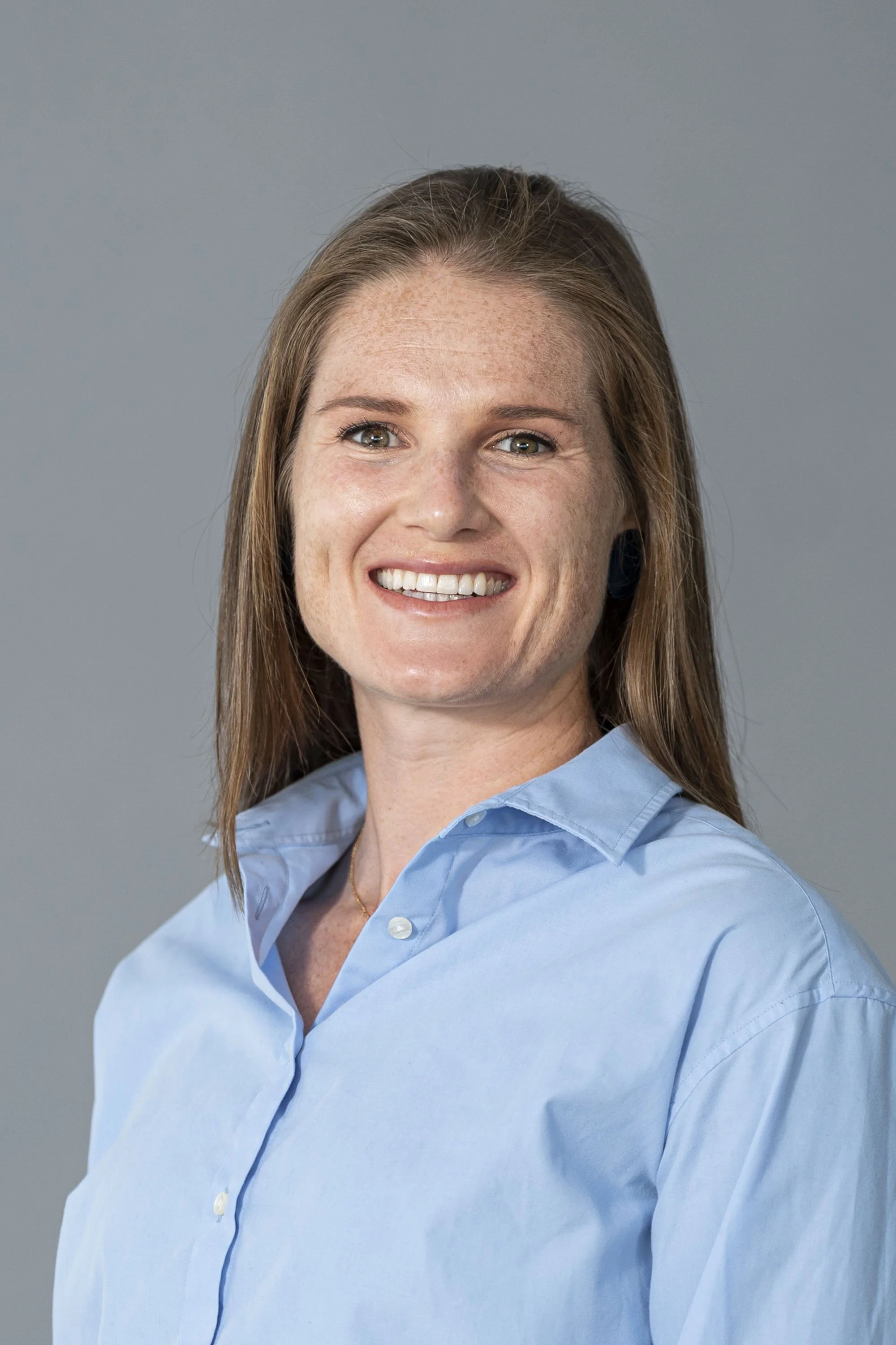 A smiling woman with long, straight brown hair, wearing a light blue collared shirt, against a gray background.