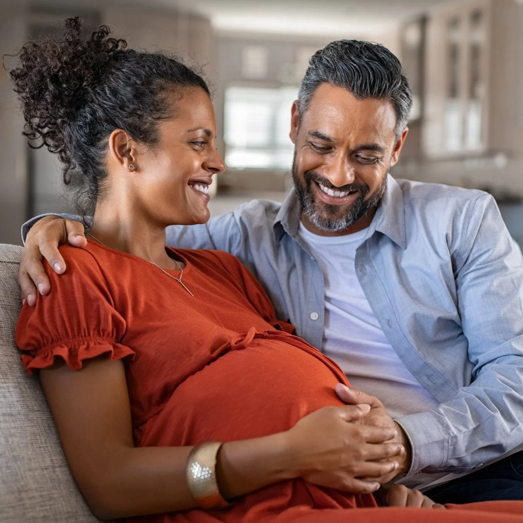 A happy pregnant woman and her partner sitting on a couch. The woman is wearing a red dress and has her hand on her belly, while the man has his arm around her shoulder and is gently touching her belly. They are smiling and looking at each other in a cozy, well-lit living room.