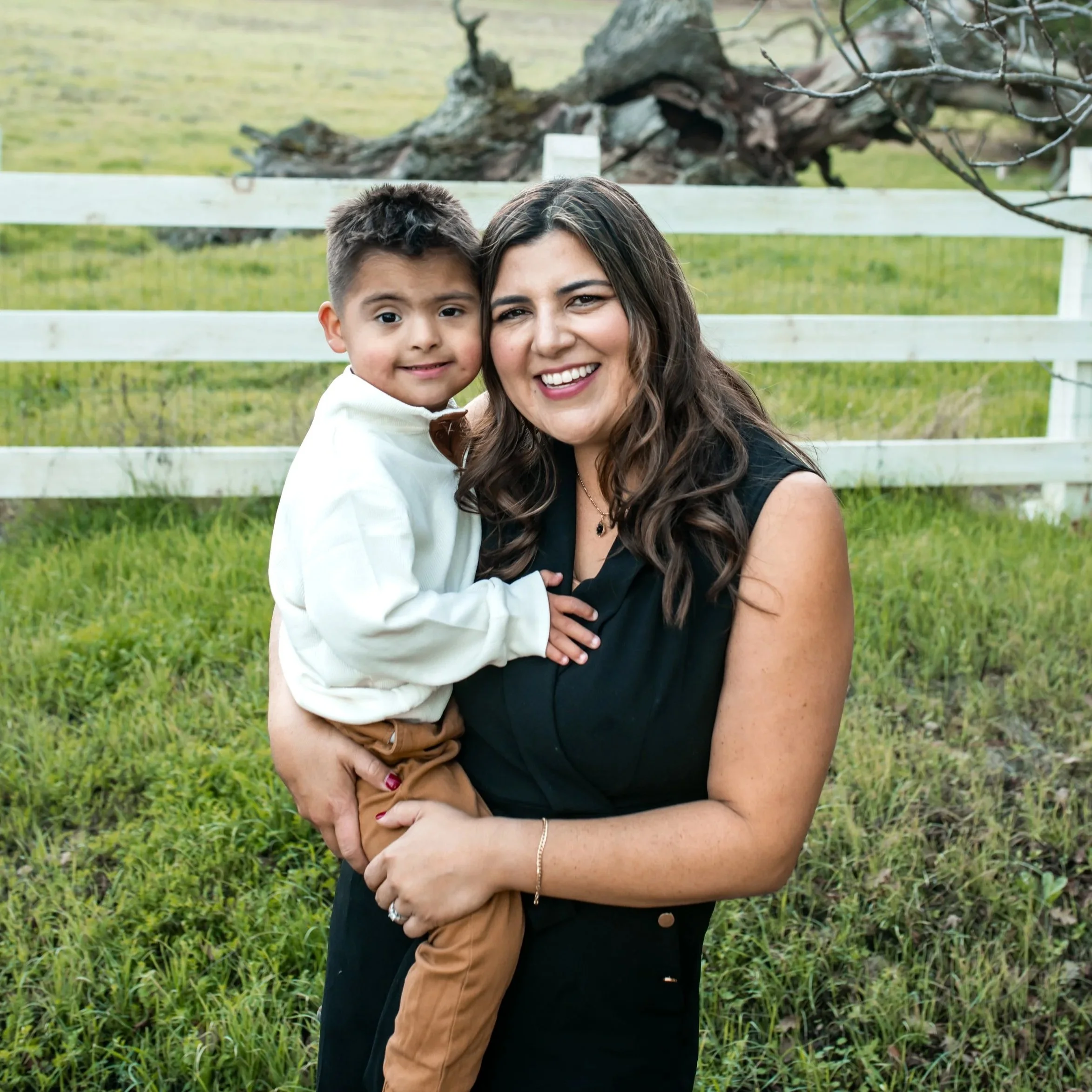 A woman and a young boy smiling and hugging outdoors in front of a white fence and a large fallen tree.