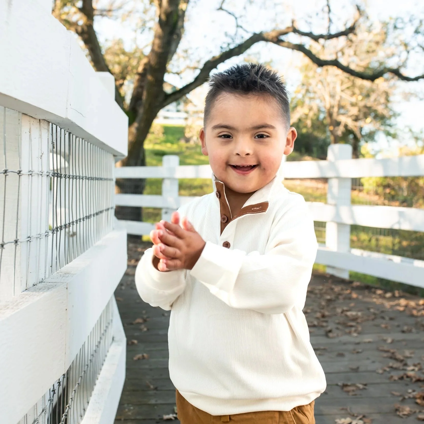A happy young boy claps his hands outdoors on a wooden bridge with a white fence, trees, and greenery in the background.