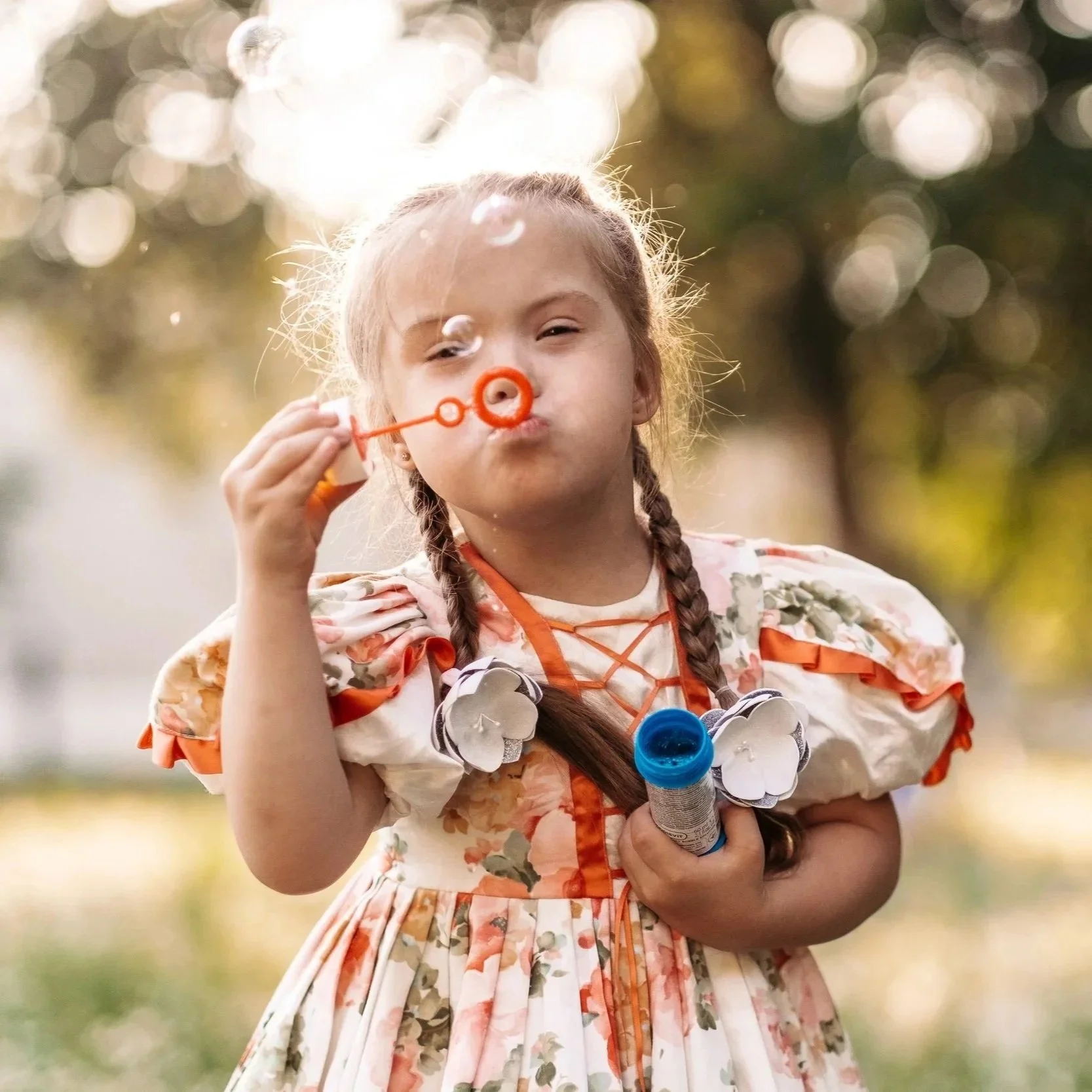 A young girl with braided hair blowing bubbles outdoors, holding a bubble wand in one hand and a container of bubble solution in the other, wearing a floral dress with ruffled sleeves.