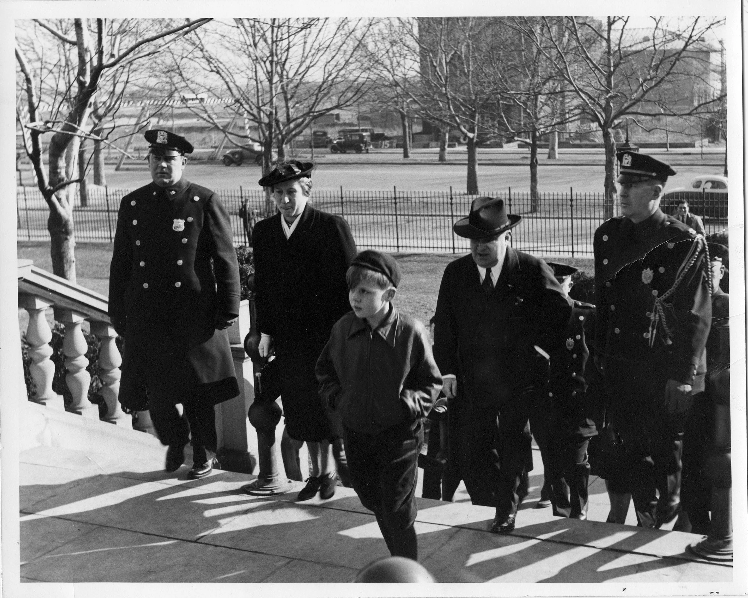 Black and white photograph of police officers and a woman walking outside in a park-like setting, with leafless trees and a fence in the background, possibly taken in the mid-20th century.