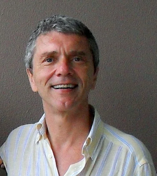 A man with gray hair smiling, wearing a light-colored, striped button-up shirt, standing against a plain dark background.