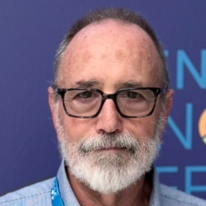 Close-up portrait of a middle-aged man with glasses, gray beard, and short gray hair, wearing a blue collared shirt and a lanyard, standing against a blue background with abstract text.