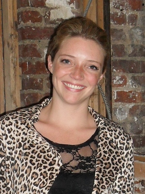 A smiling woman with short, light brown hair wearing a leopard print jacket over a black lace top, standing in front of a brick wall with exposed wooden framing.