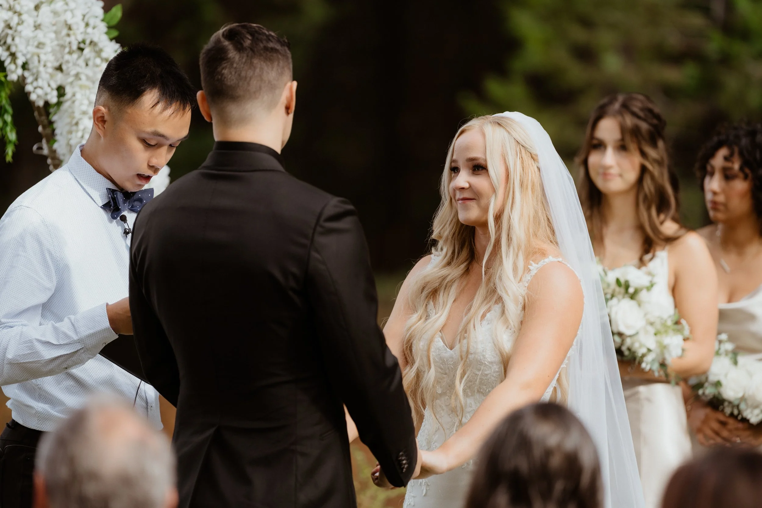 A bride and groom exchanging vows during their outdoor wedding ceremony, with bridesmaids holding bouquets in the background.