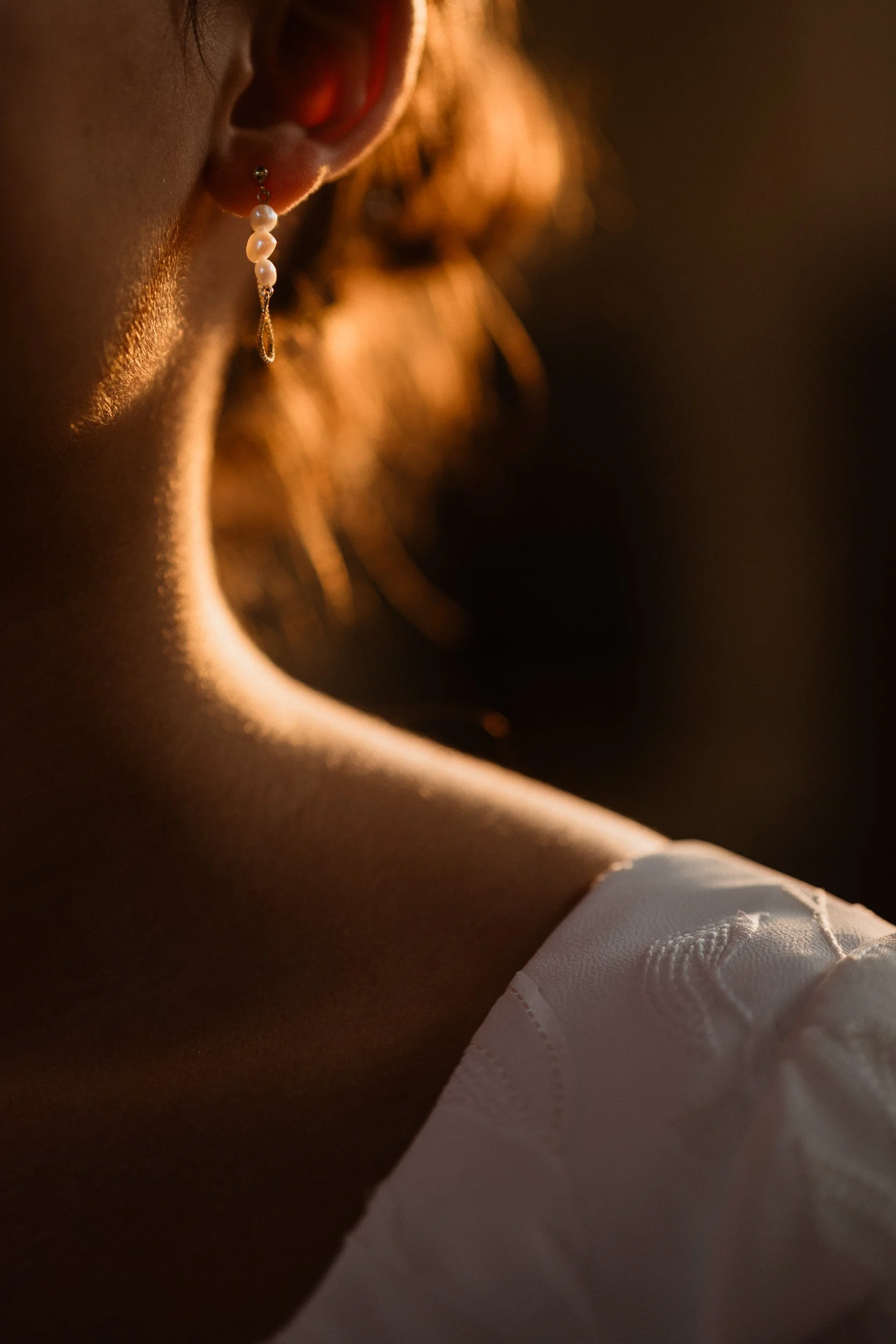 Close-up of a woman's ear with a dangling pearl earring, illuminated by warm sunlight, with a white embroidered garment visible at the bottom.