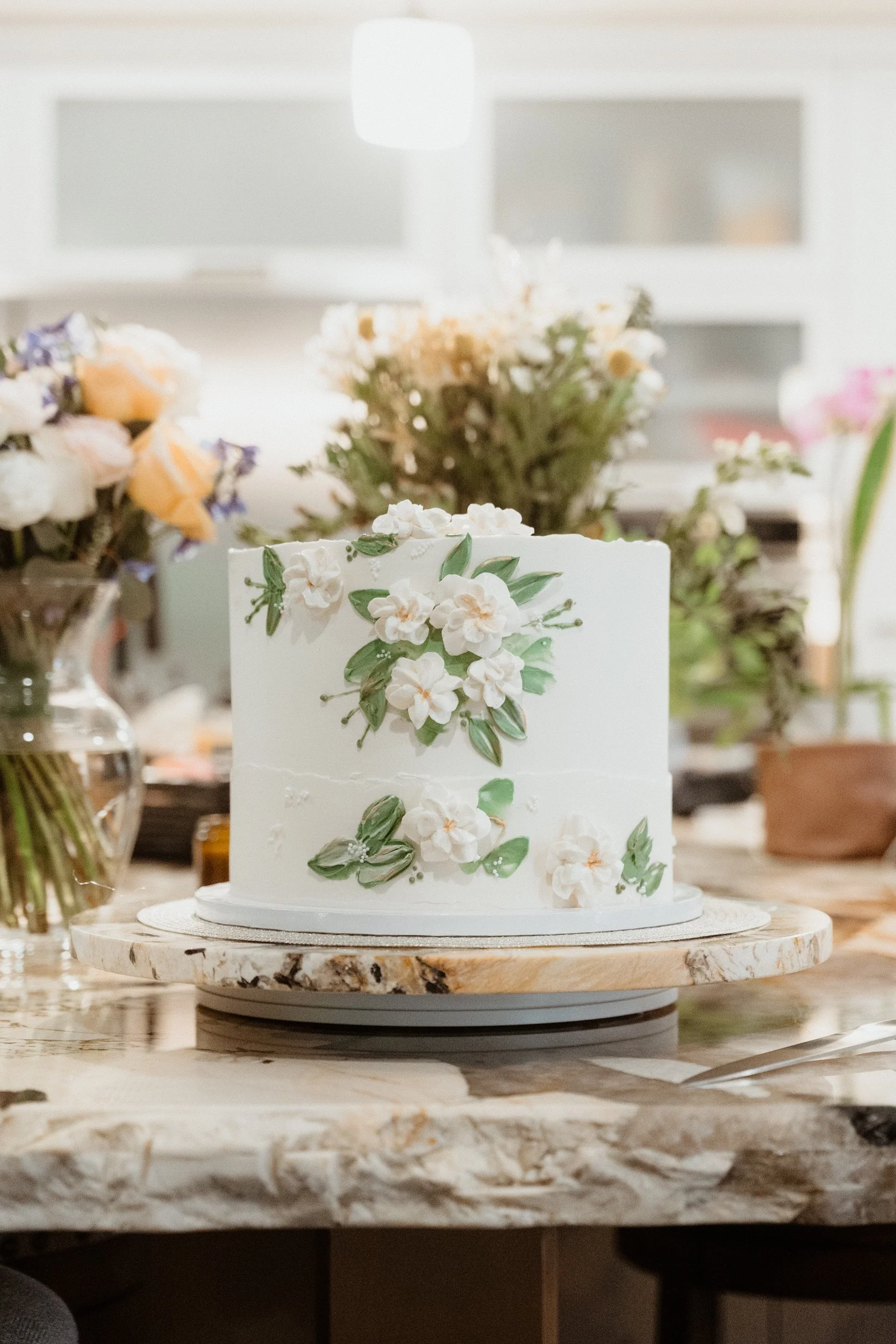 A two-tier white wedding cake decorated with white flowers and green leaves, placed on a round marble cake stand on a wooden table with flower arrangements in vases around it.