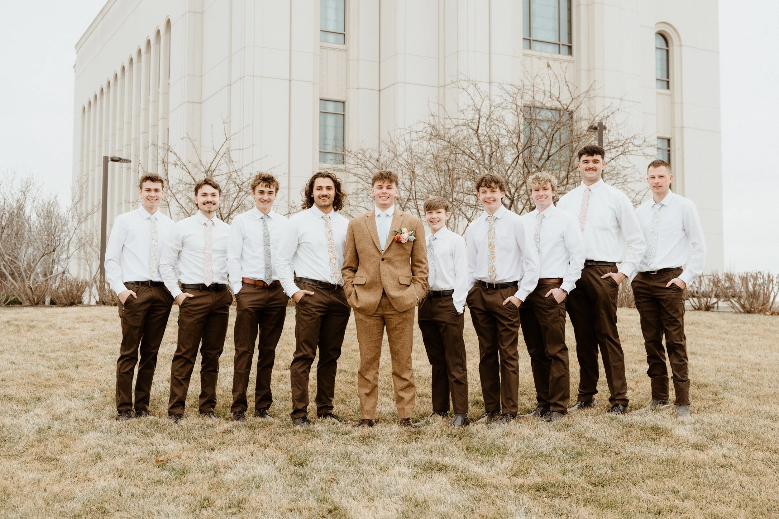 A group of eleven men standing outdoors in front of a white building, dressed in white shirts and brown pants, with the groom in a brown suit with a boutonniere, posing for a wedding photo.