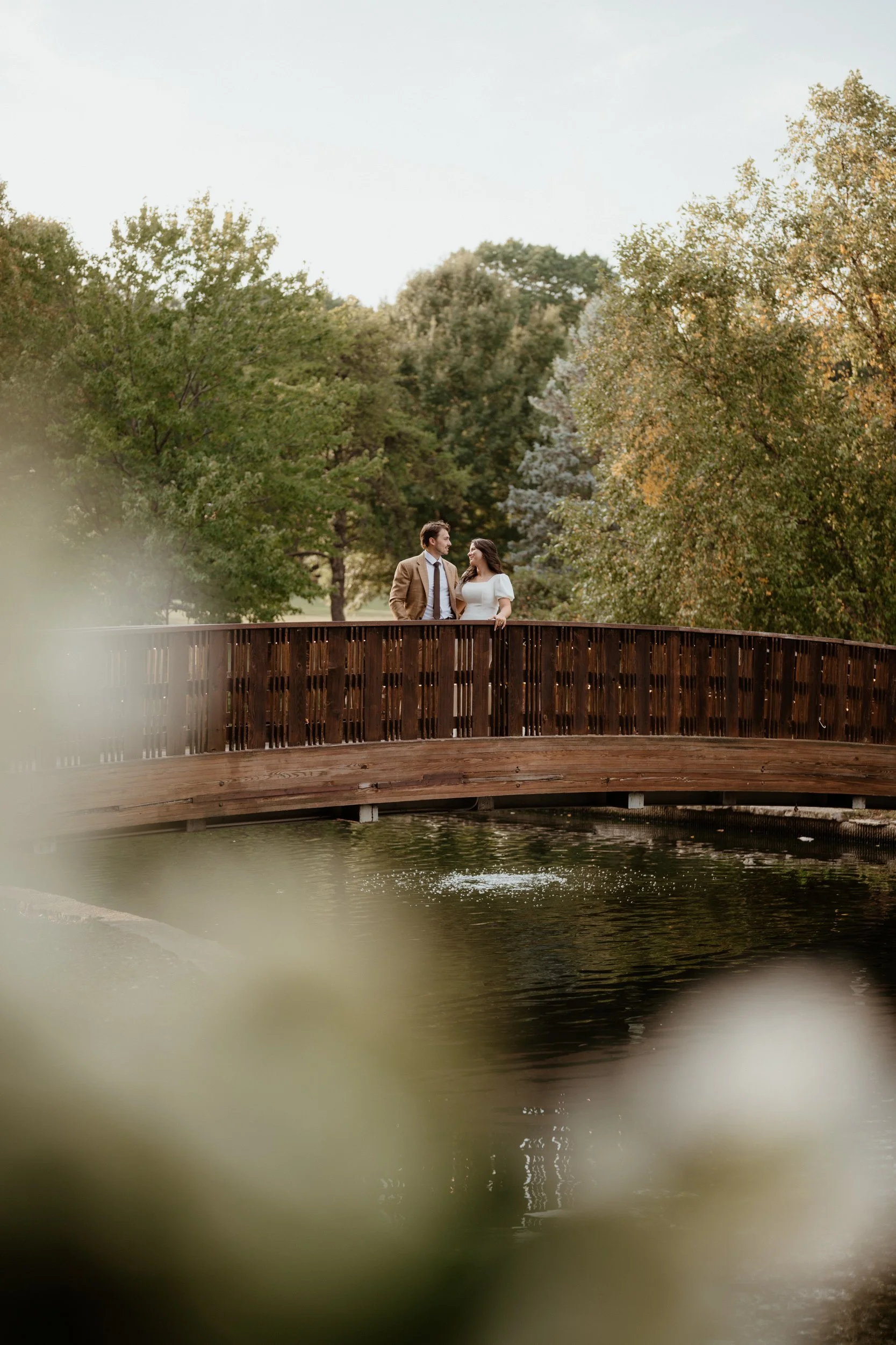 A couple standing on a wooden bridge over a pond, surrounded by lush green trees during late afternoon or early evening.