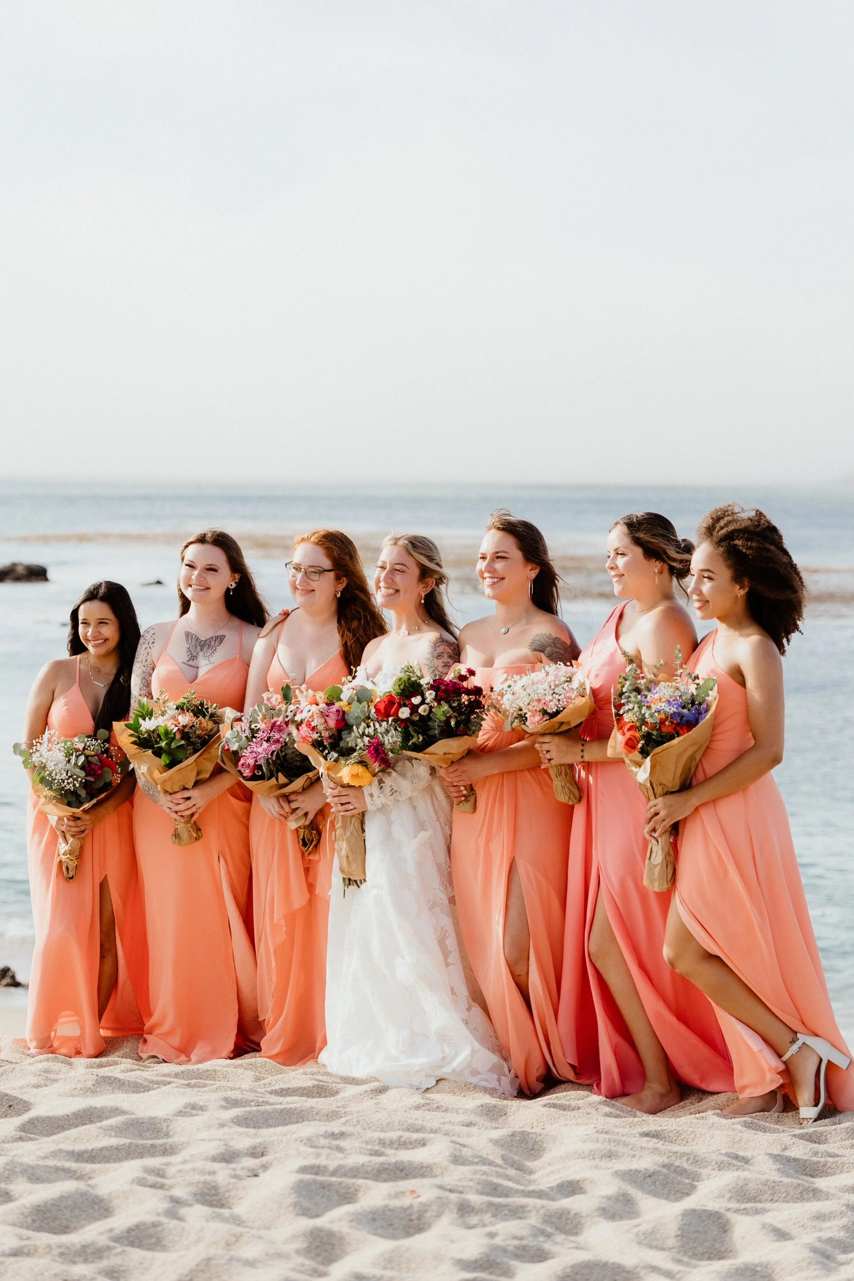 Bride and bridesmaids in peach dresses standing on the beach holding bouquets, smiling.