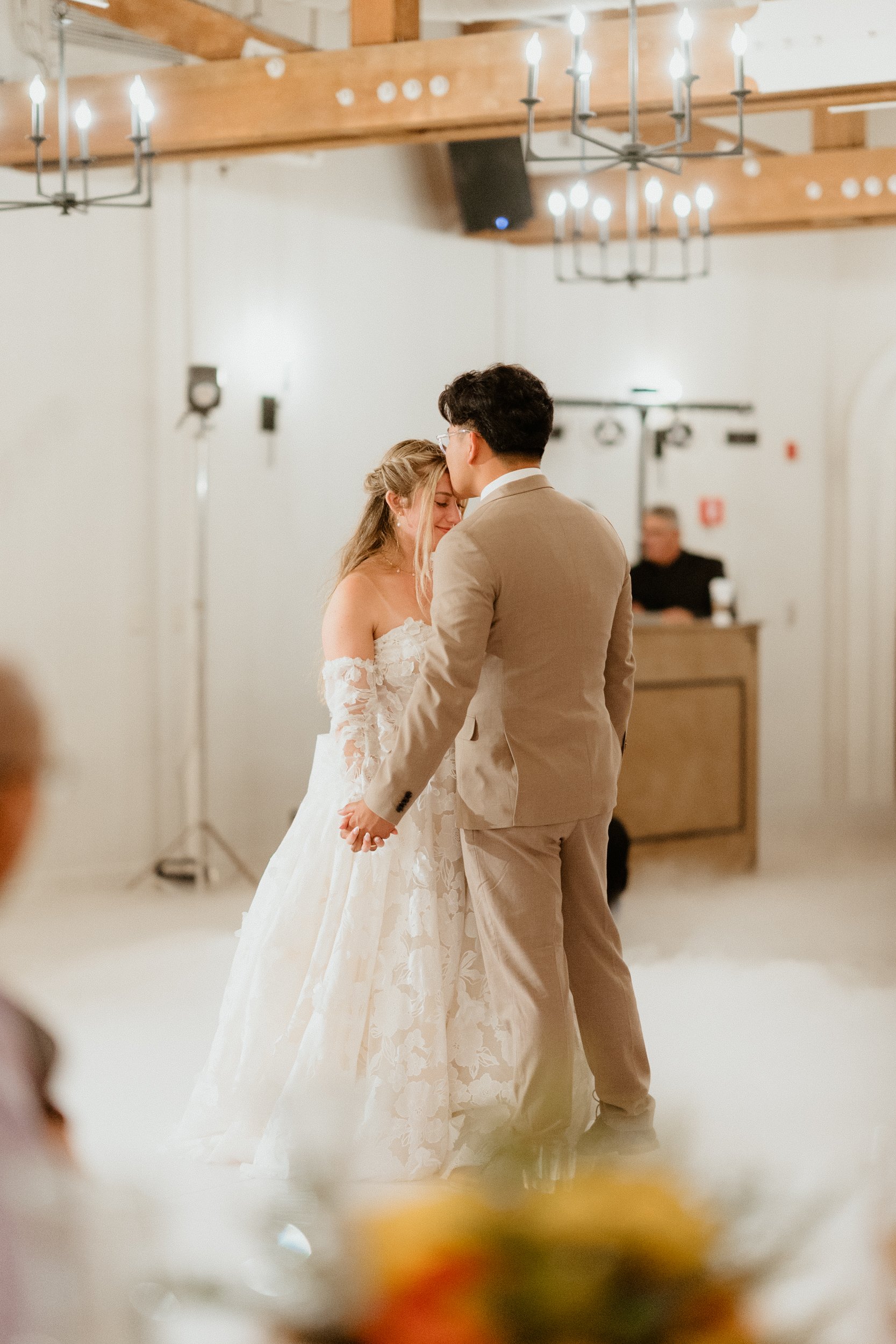 A bride and groom sharing their first dance at a wedding reception, holding hands and embracing in a warmly lit venue with chandeliers and wooden beams.