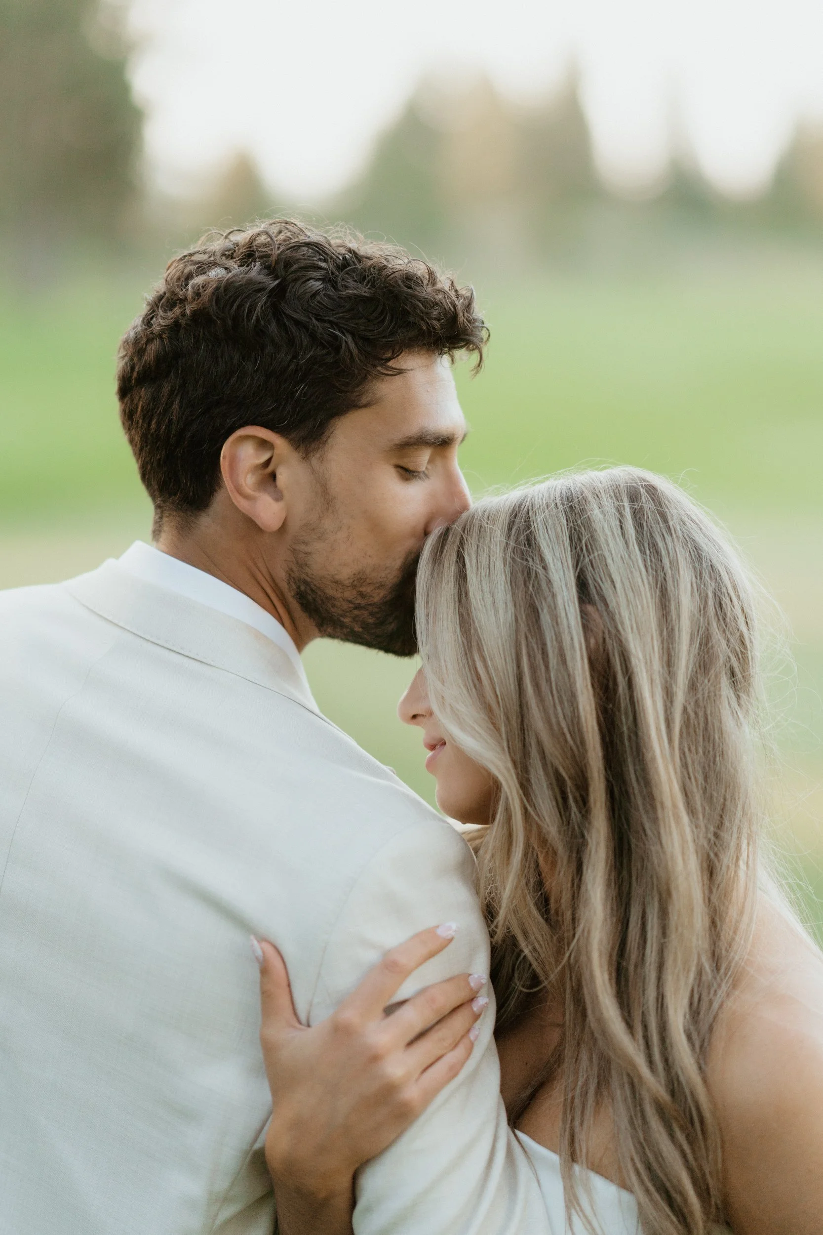 A couple hugging outdoors, the man is kissing the woman on the forehead, with a blurred green landscape in the background.