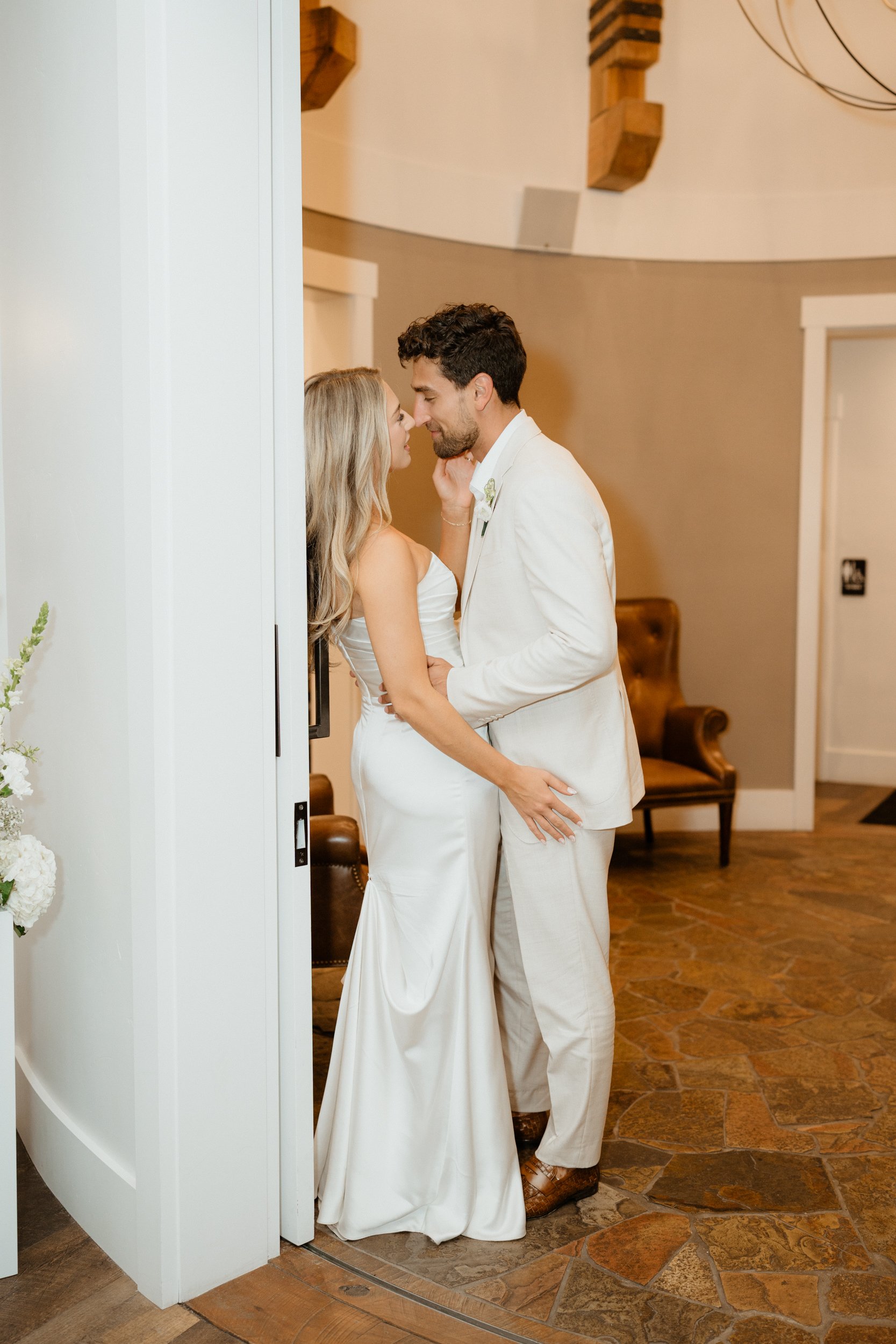 A bride and groom are close together inside a room, sharing an intimate moment. The bride is wearing a strapless white wedding dress, and the groom is dressed in a white suit. They are touching foreheads and gazing into each other's eyes.