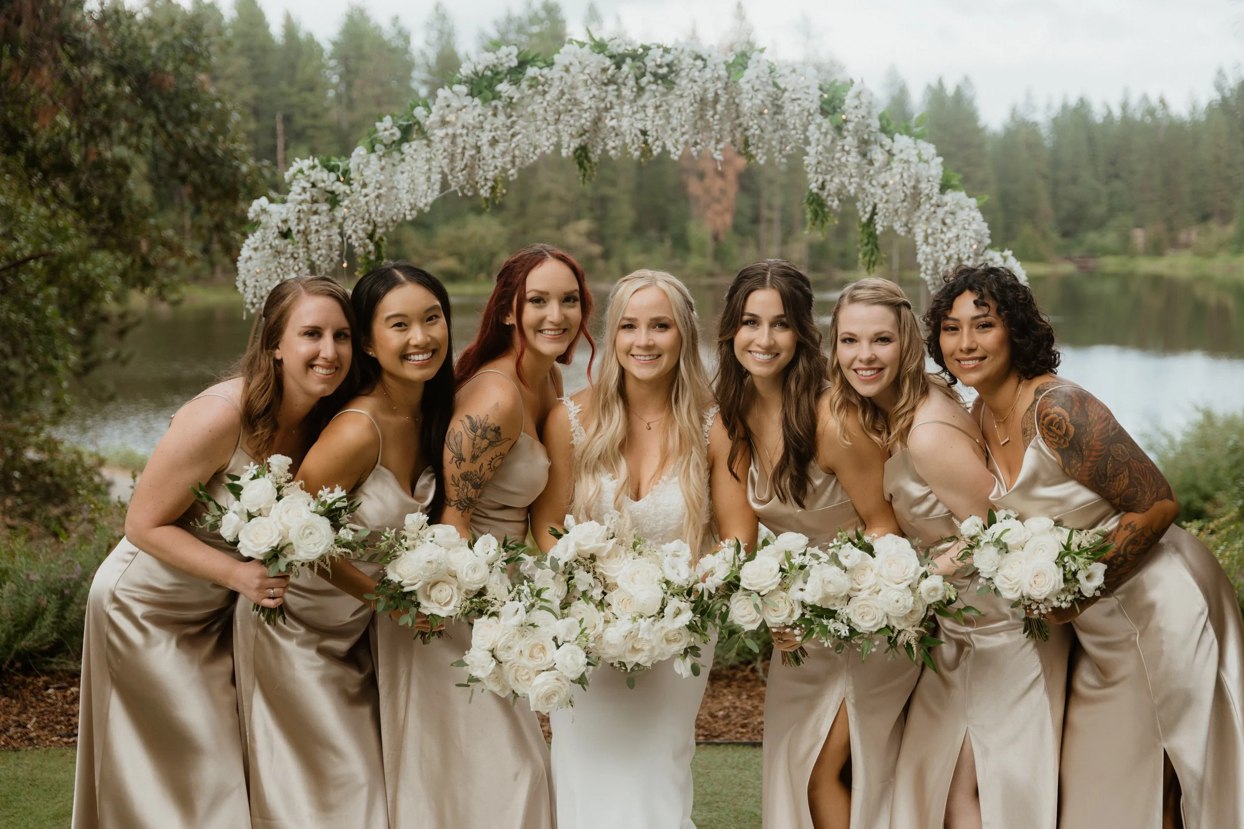 A bride and six bridesmaids standing outdoors near a lake, smiling, holding white floral bouquets, with a floral arch behind them.