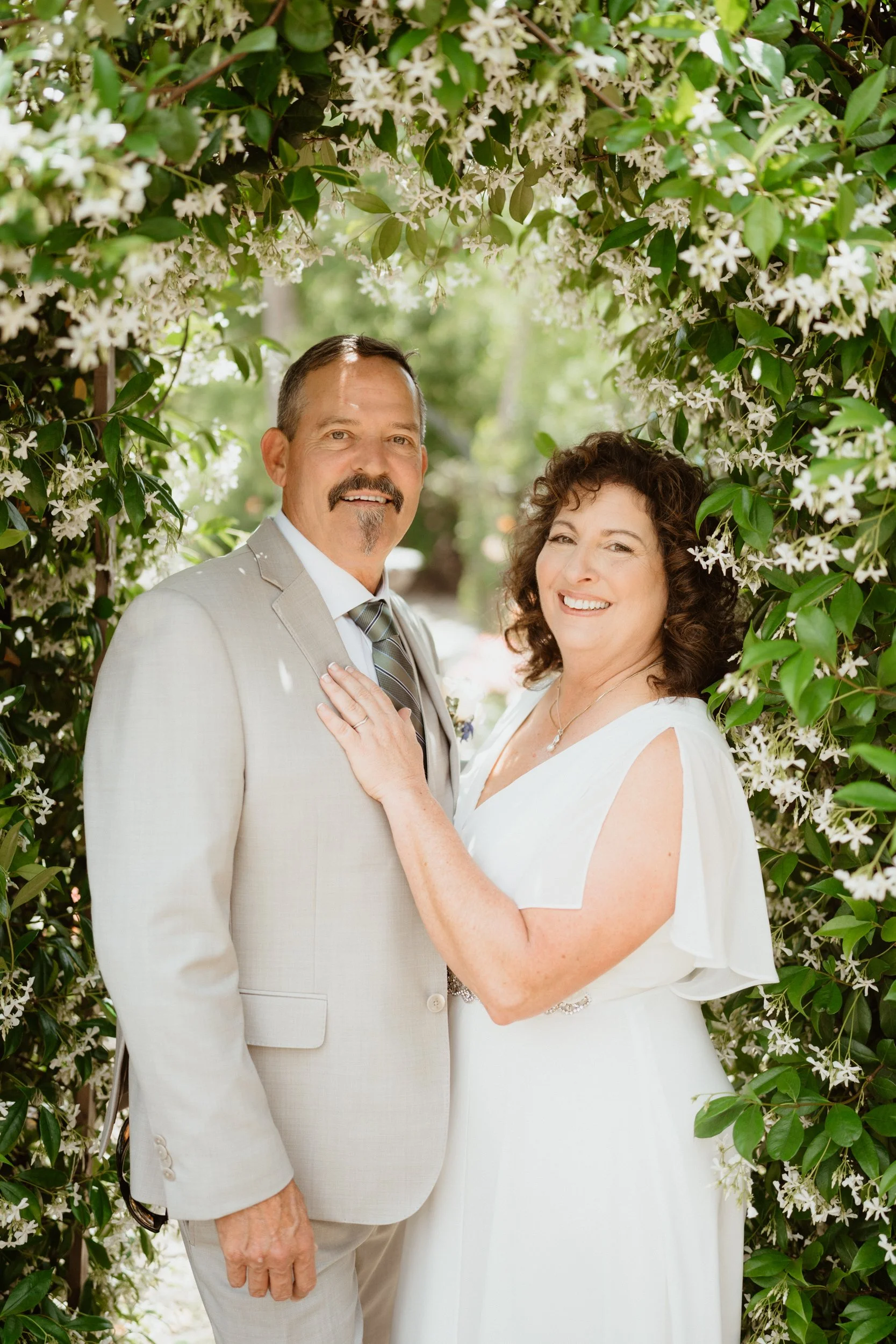 A happy couple on their wedding day, standing in a floral archway outdoors, smiling at the camera.